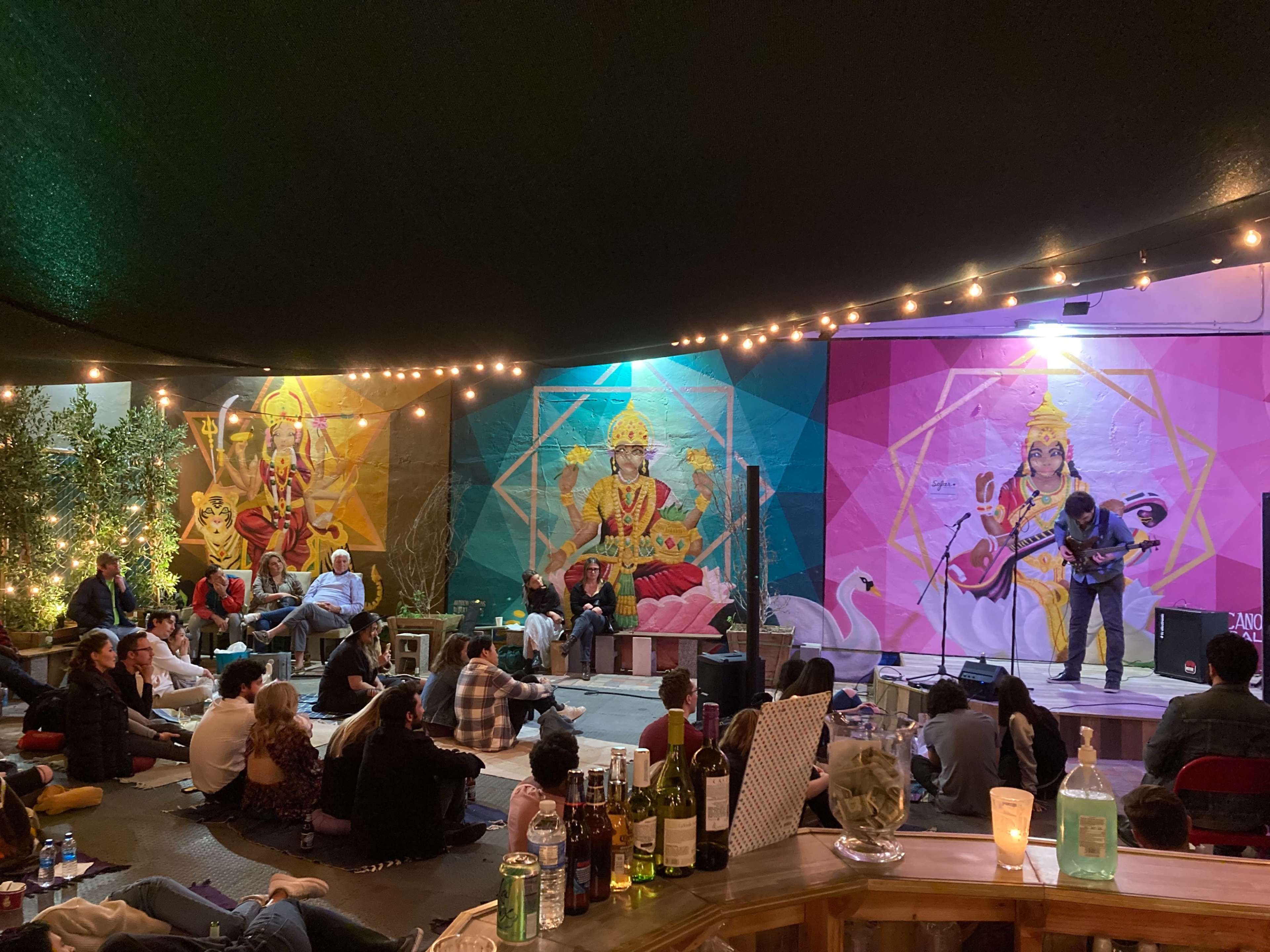 A musician performs on stage while an audience listens, surrounded by colorful murals and string lights in an outdoor space.