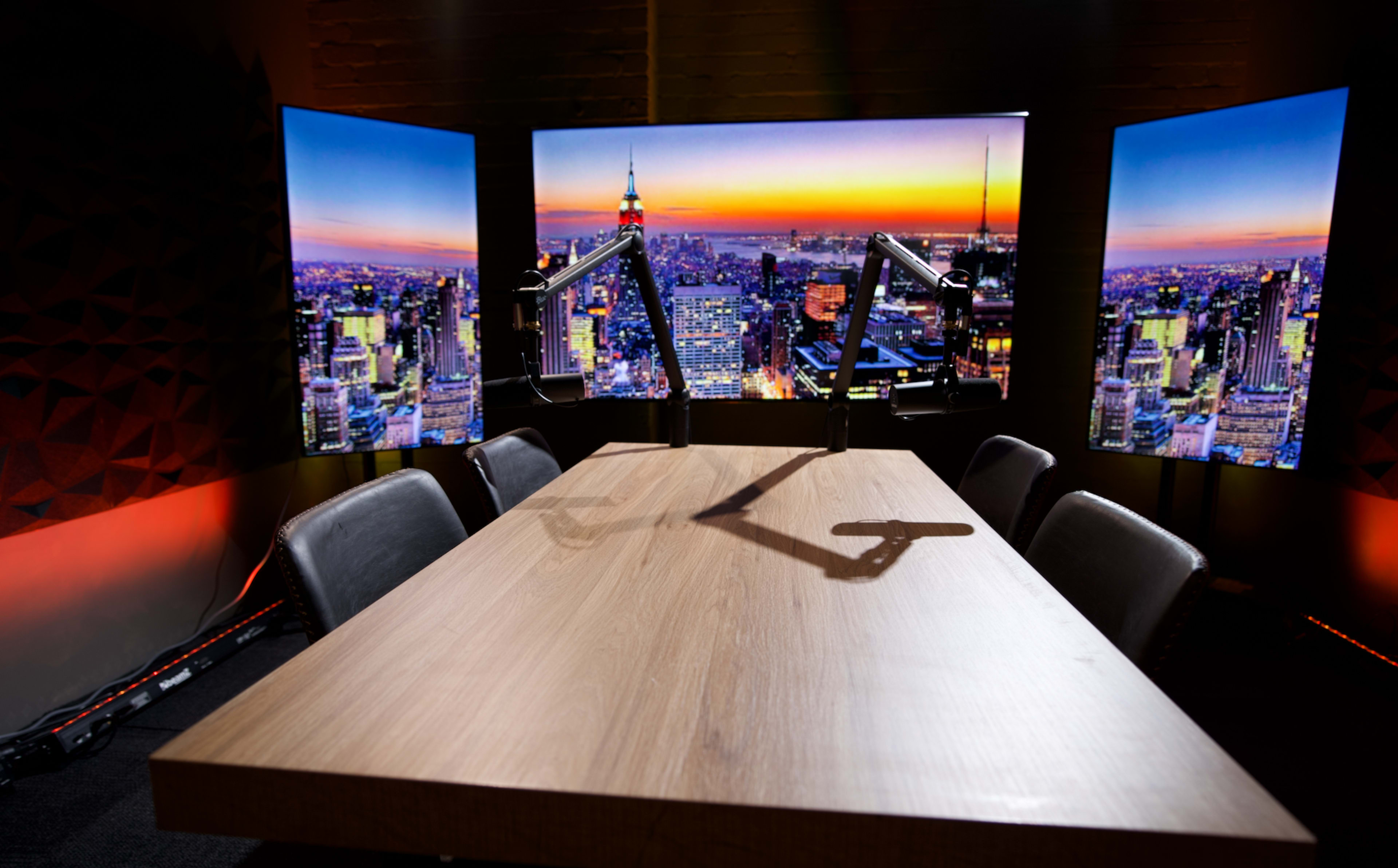 A modern conference room is set up with a large wooden table, equipped with microphone arms, and three screens displaying a city skyline at sunset.