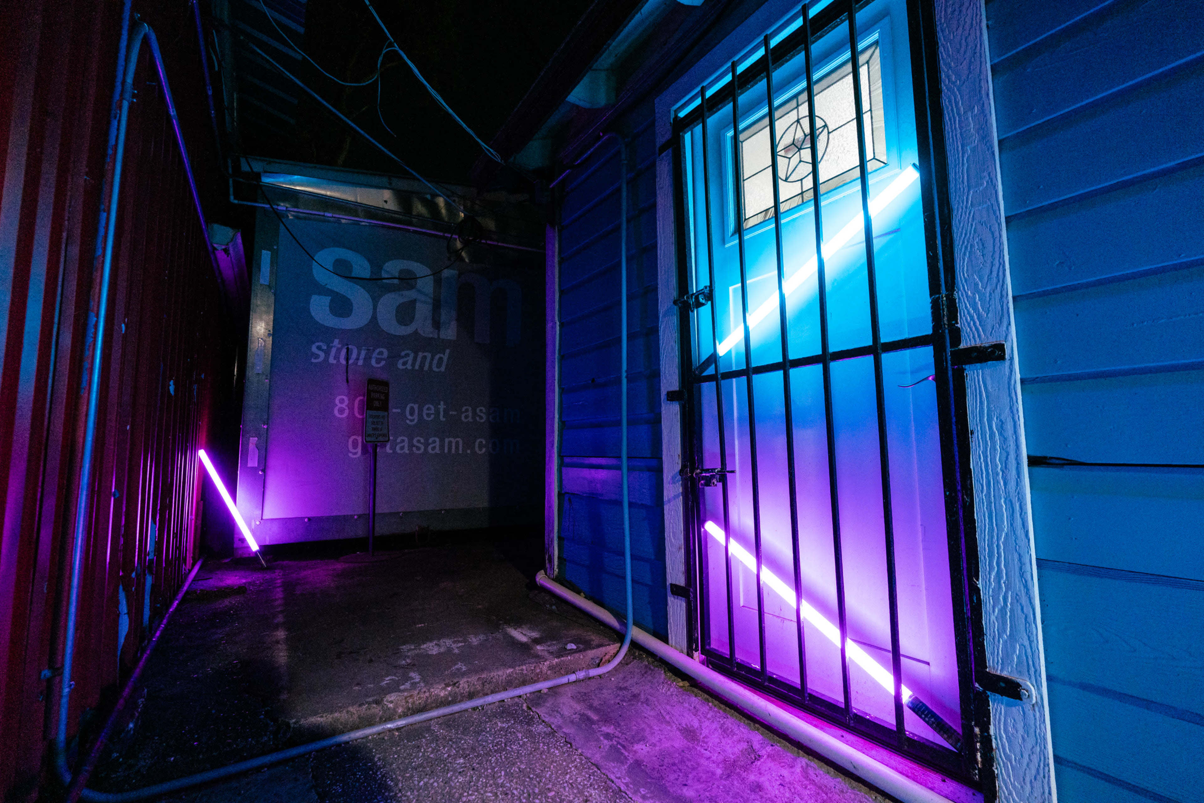 A narrow alleyway with a blue wooden door secured by a metal grate, illuminated by neon purple lights.