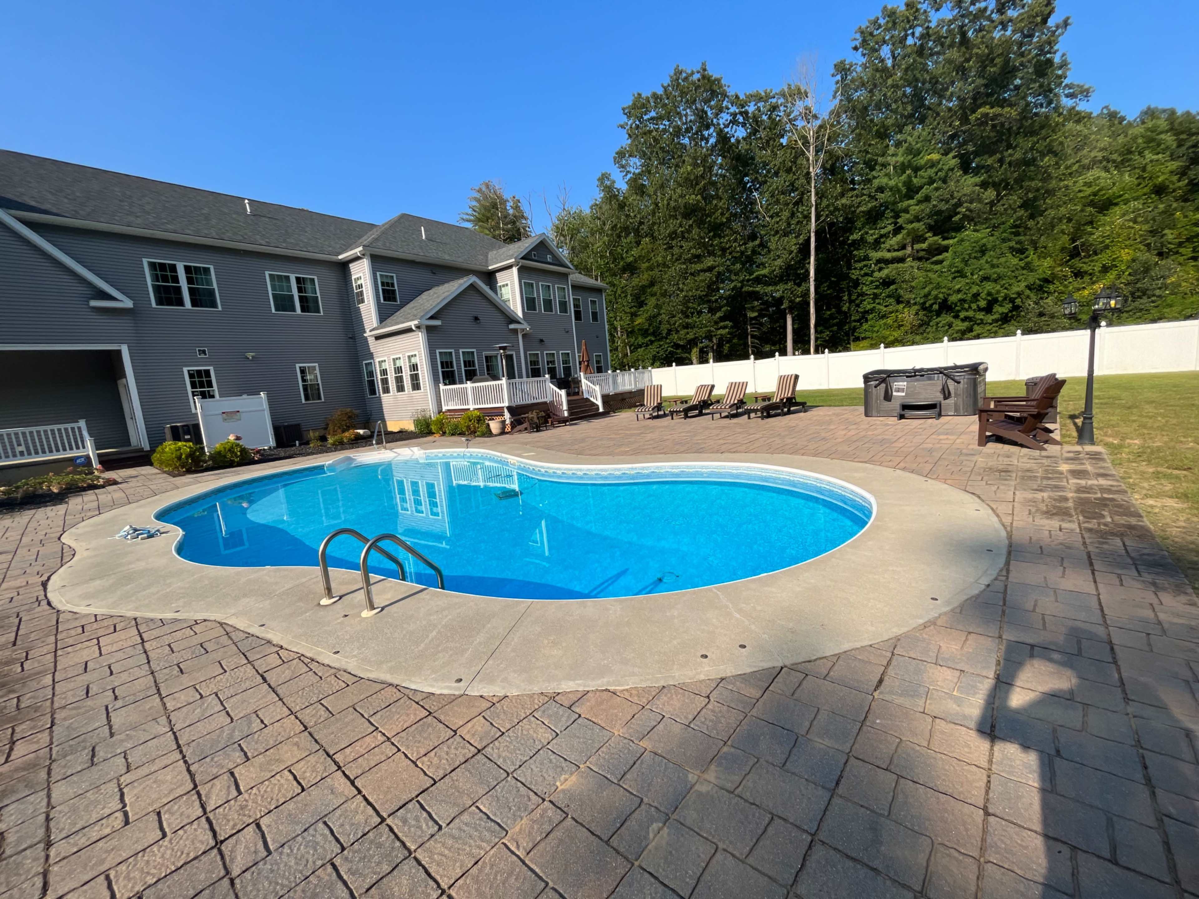 A residential backyard with a blue swimming pool surrounded by a stone patio and several lounge chairs, with a large house and trees in the background.