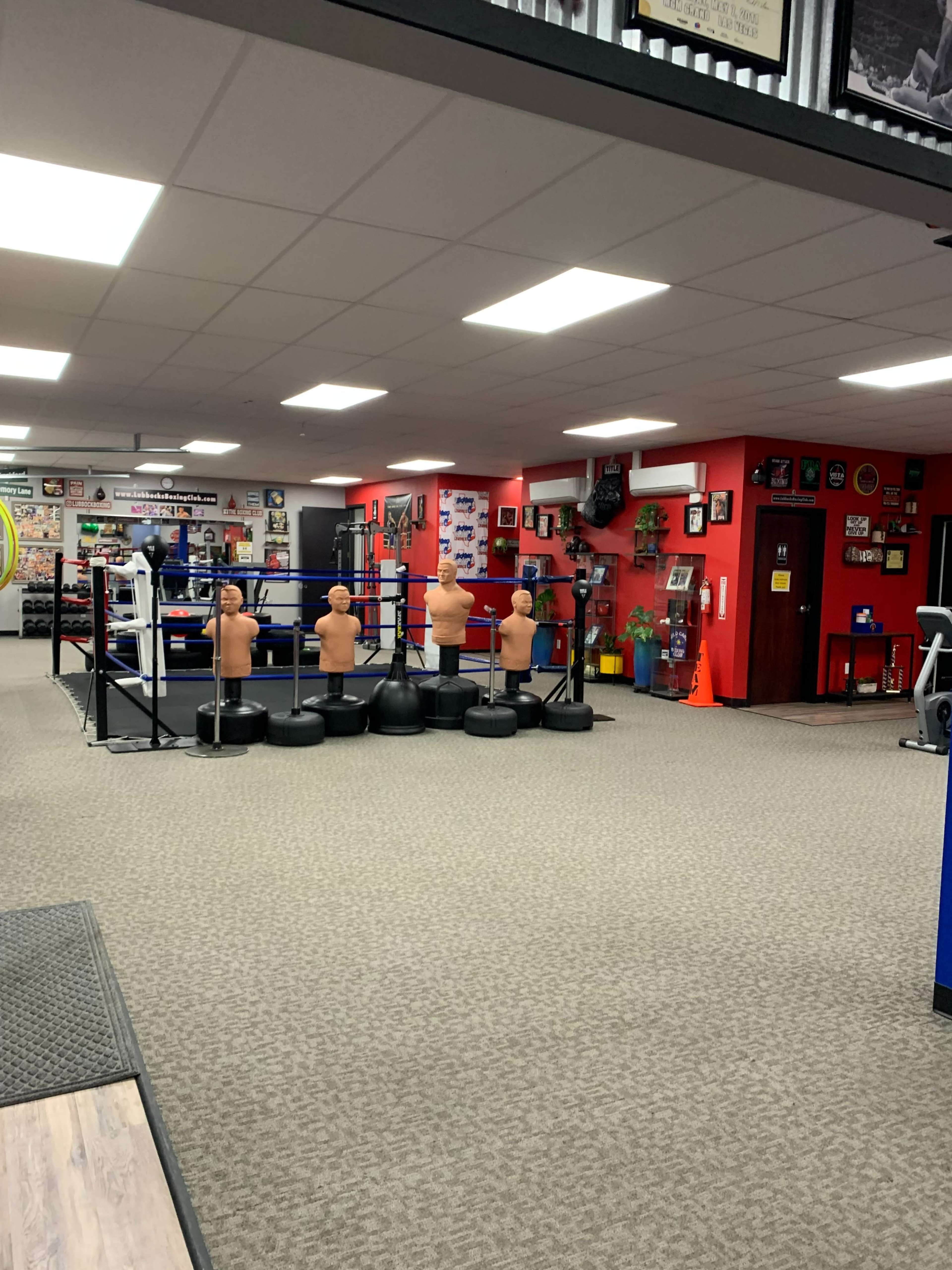 An empty gym featuring a boxing ring, several training dummies, and a variety of fitness equipment against a backdrop of colorful walls adorned with pictures and memorabilia.