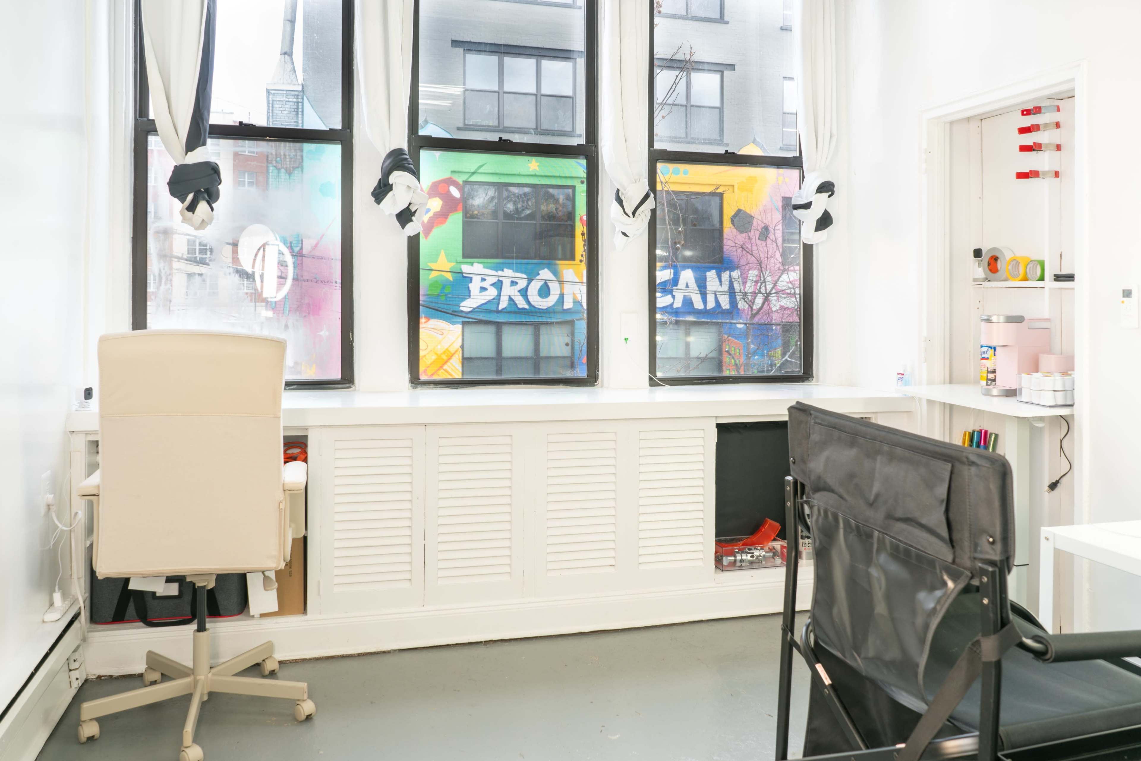 A bright room with two windows displaying colorful graffiti on the building outside, featuring the word "BRONX" and other designs, along with a white desk and black chair.