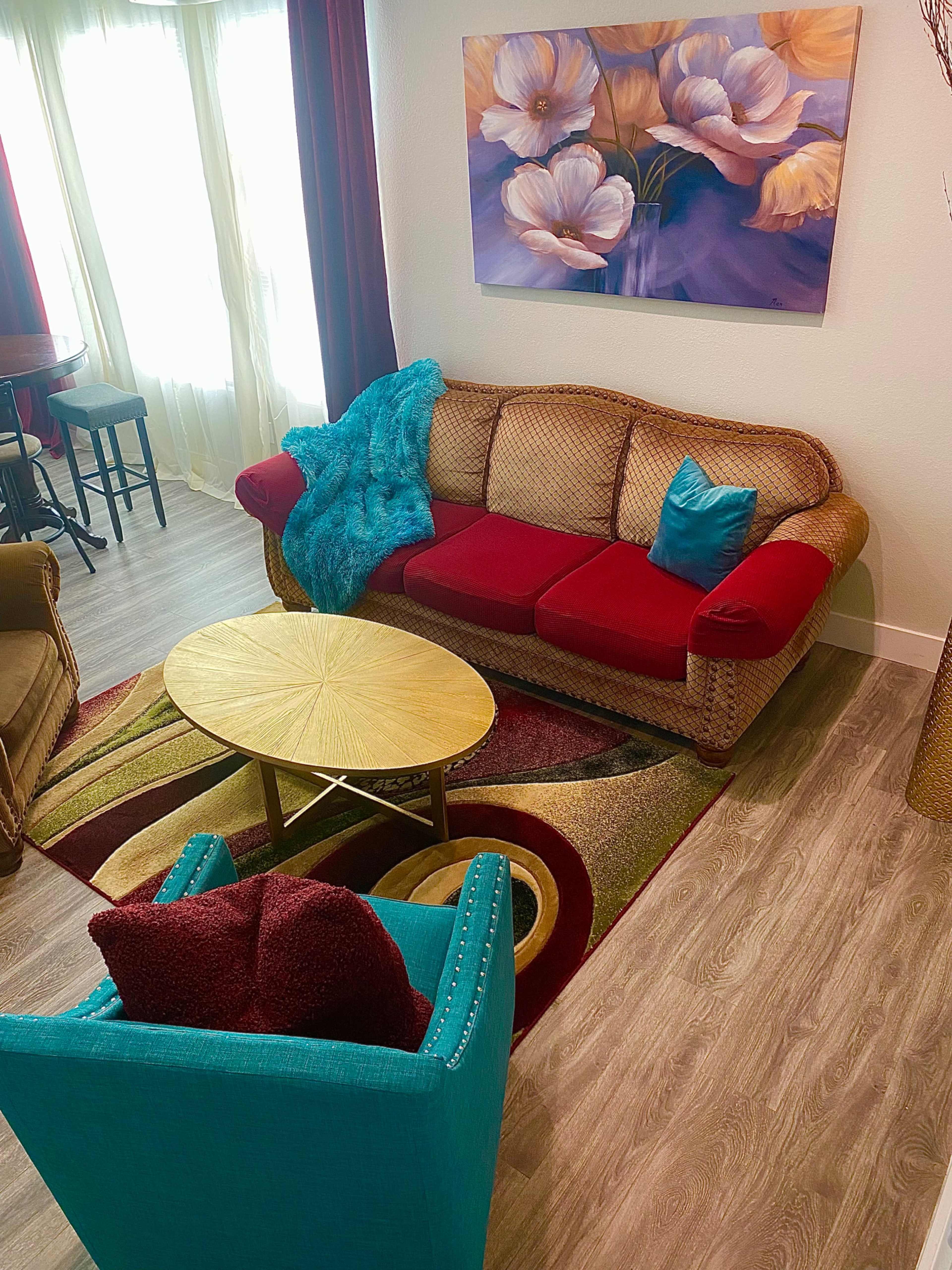 The image shows a living room with a red and beige sofa, a round gold coffee table, a turquoise armchair, and a floral painting on the wall.