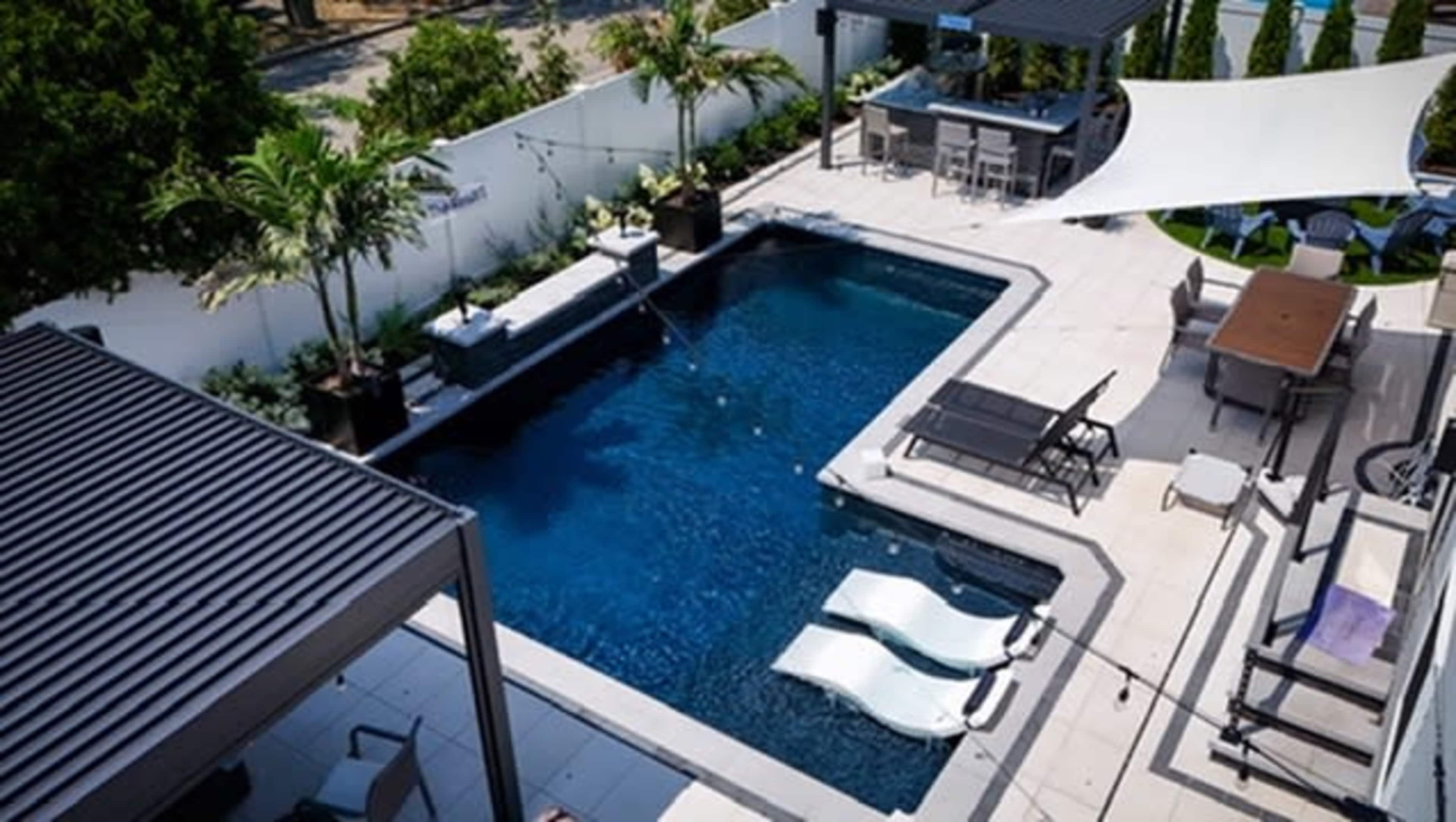 The image shows a modern backyard featuring a rectangular swimming pool surrounded by lounge chairs, a dining area, and shaded structures.