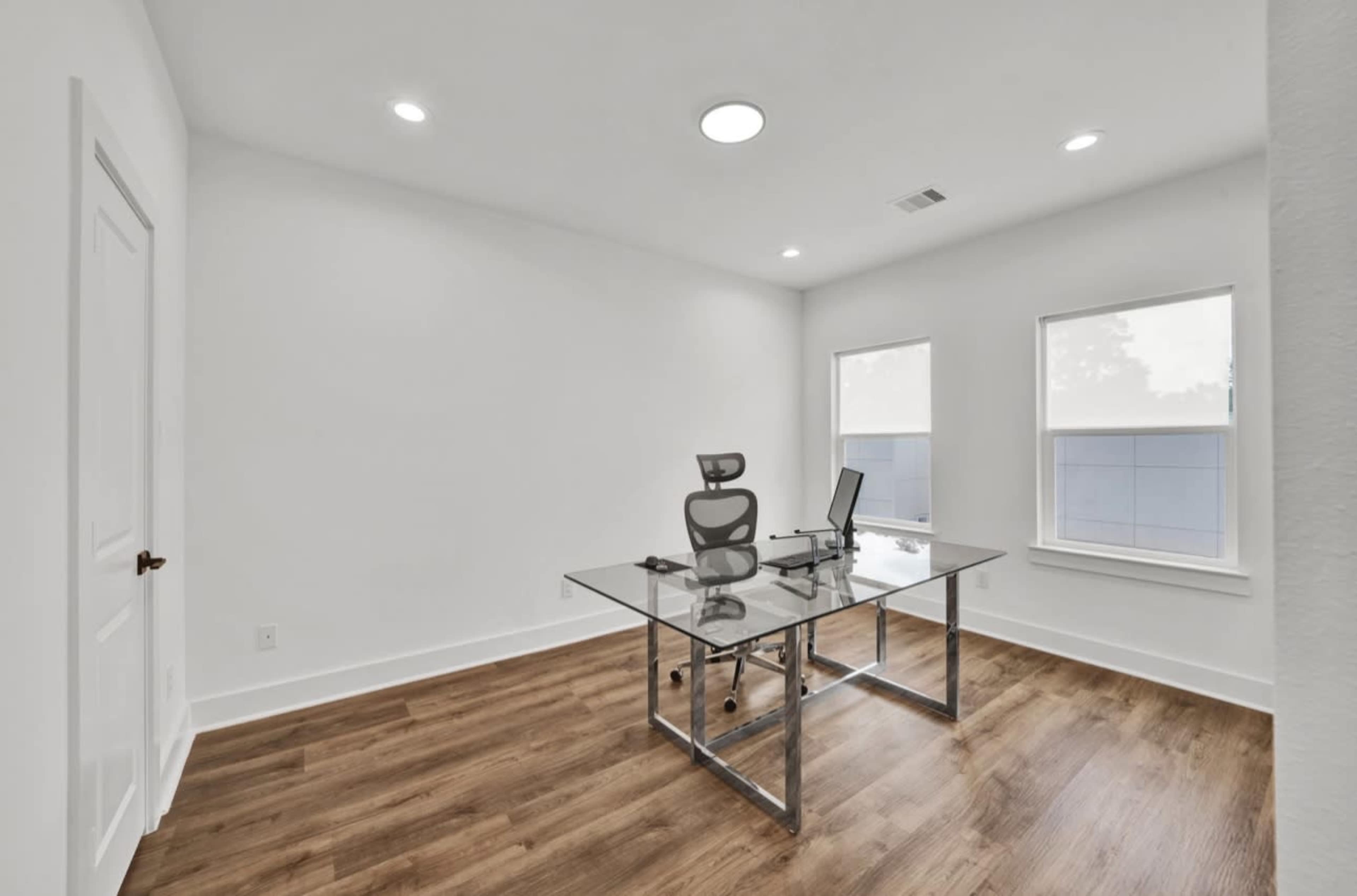 The image shows a modern office space featuring a glass desk, a swivel chair, and large windows that allow natural light to enter, with light-colored walls and wooden flooring.