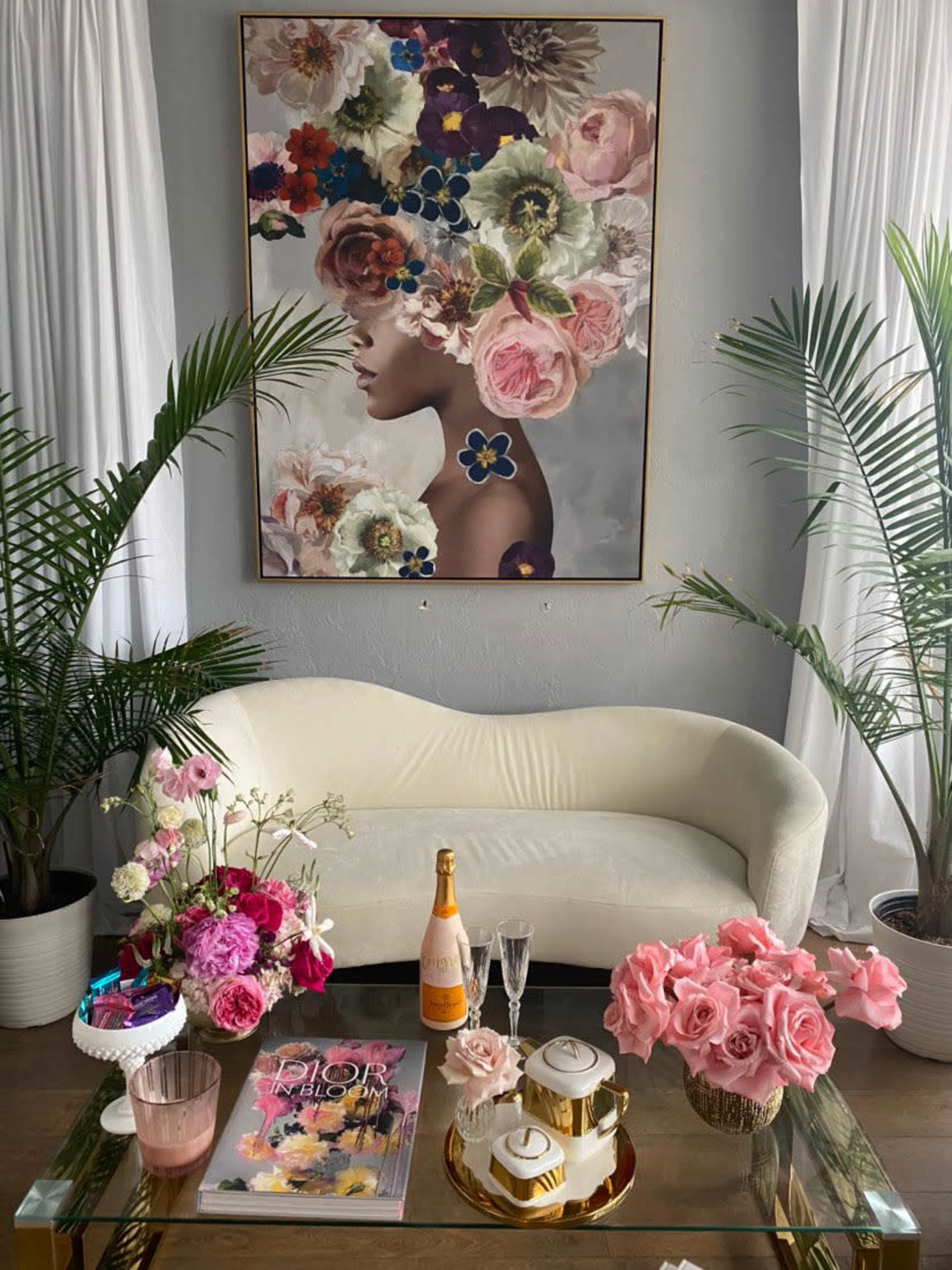 A stylish living room features a floral-themed painting above a pale couch, with a coffee table adorned with flowers, a magazine, and a bottle of champagne.