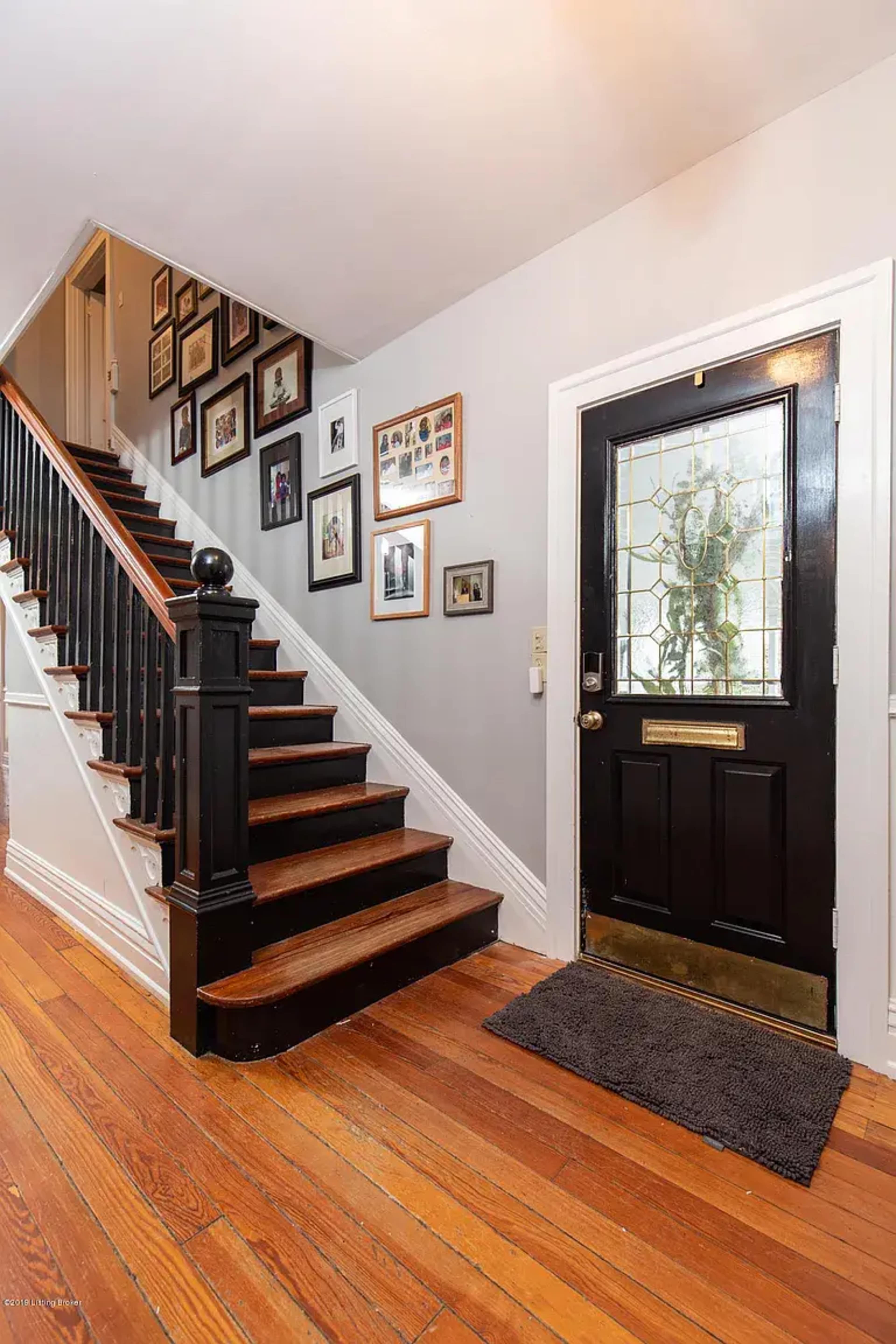 The scene shows a hallway with a staircase leading to the upper floor, framed photographs on the walls, and a front door with glass panels.