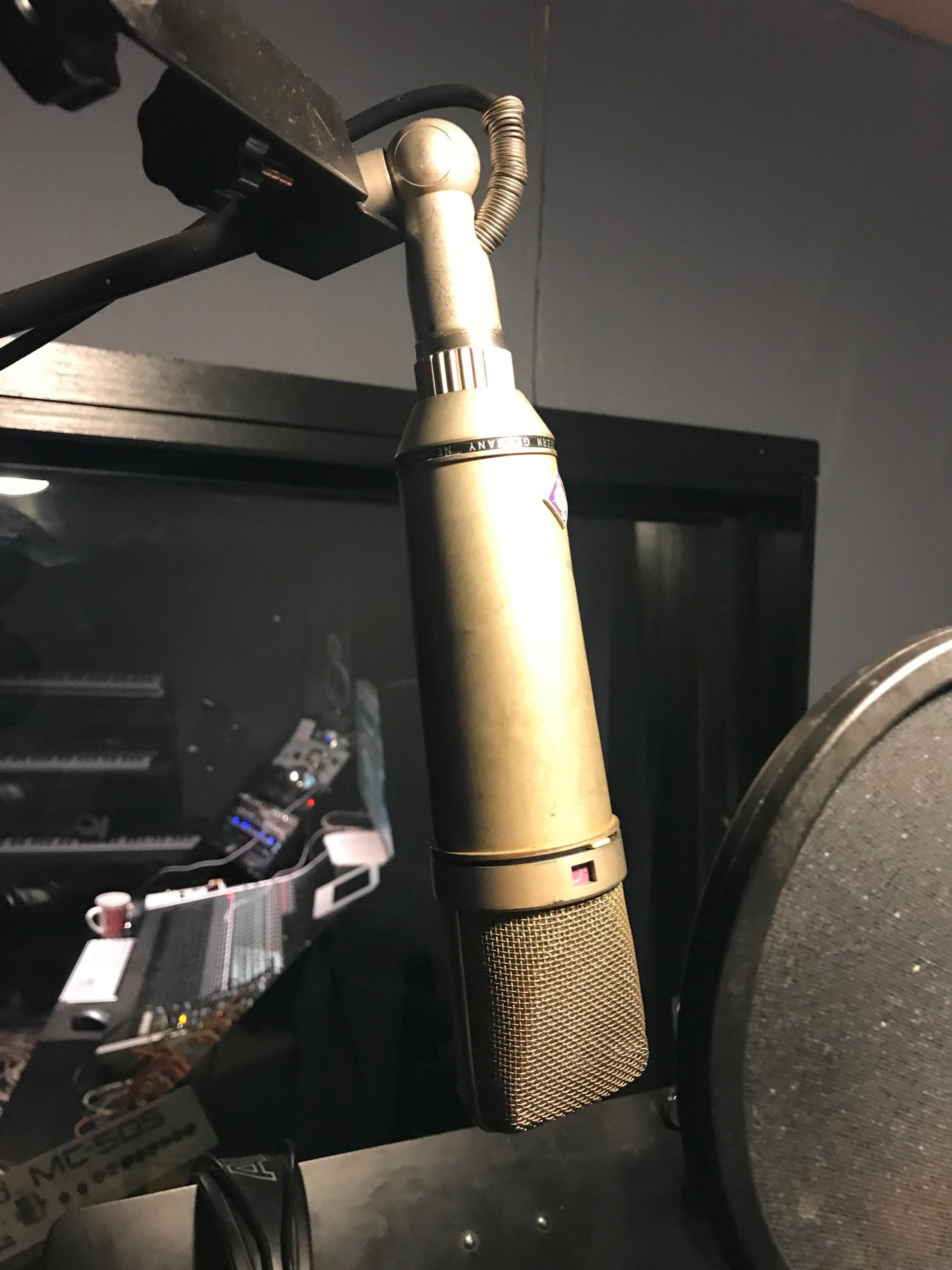 A vintage microphone is mounted on a microphone stand in a recording studio, with equipment and a console visible in the background.