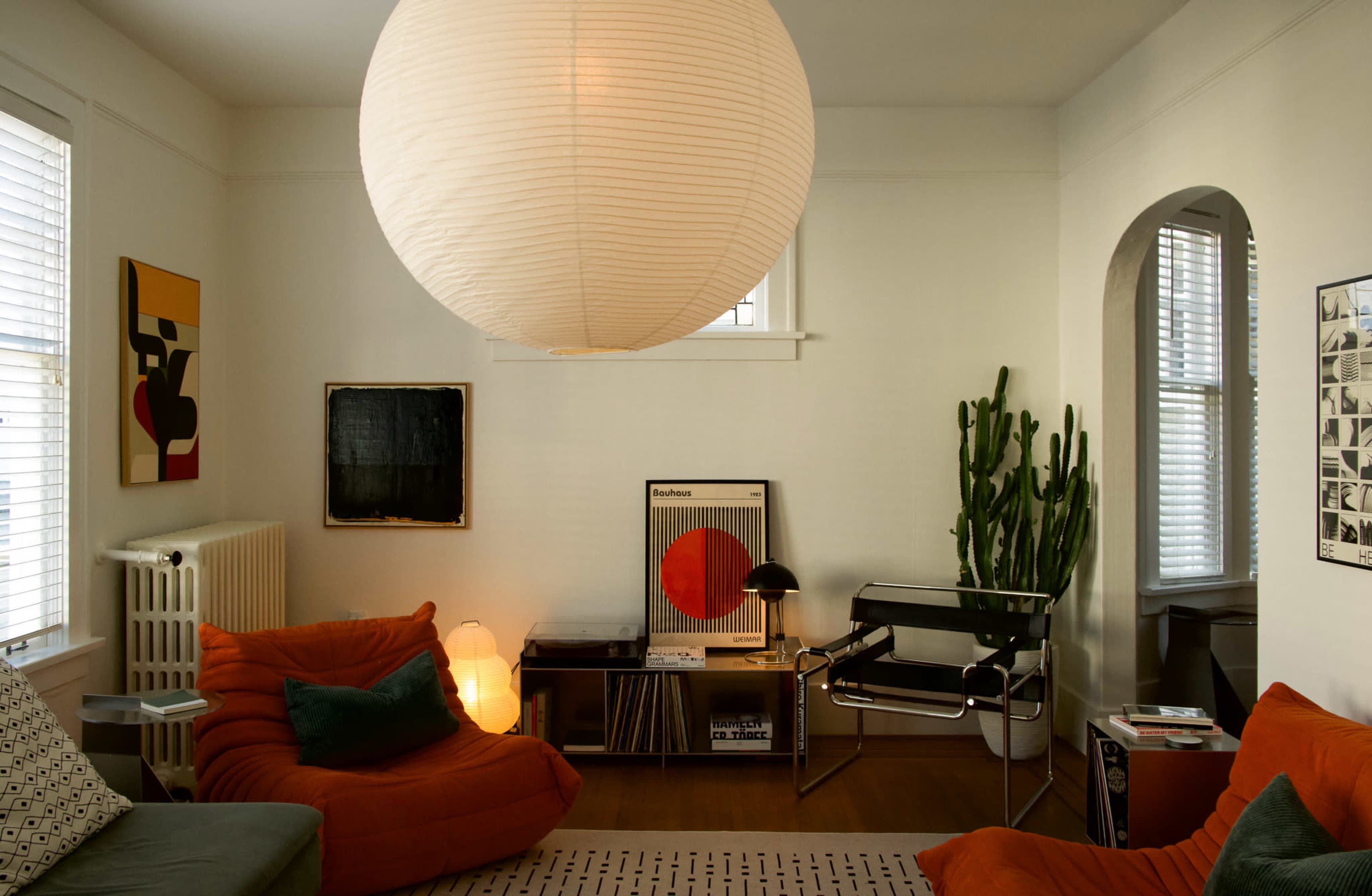 The image shows a modern living room featuring orange floor cushions, a large round pendant light, a cactus, and various artwork on the walls.