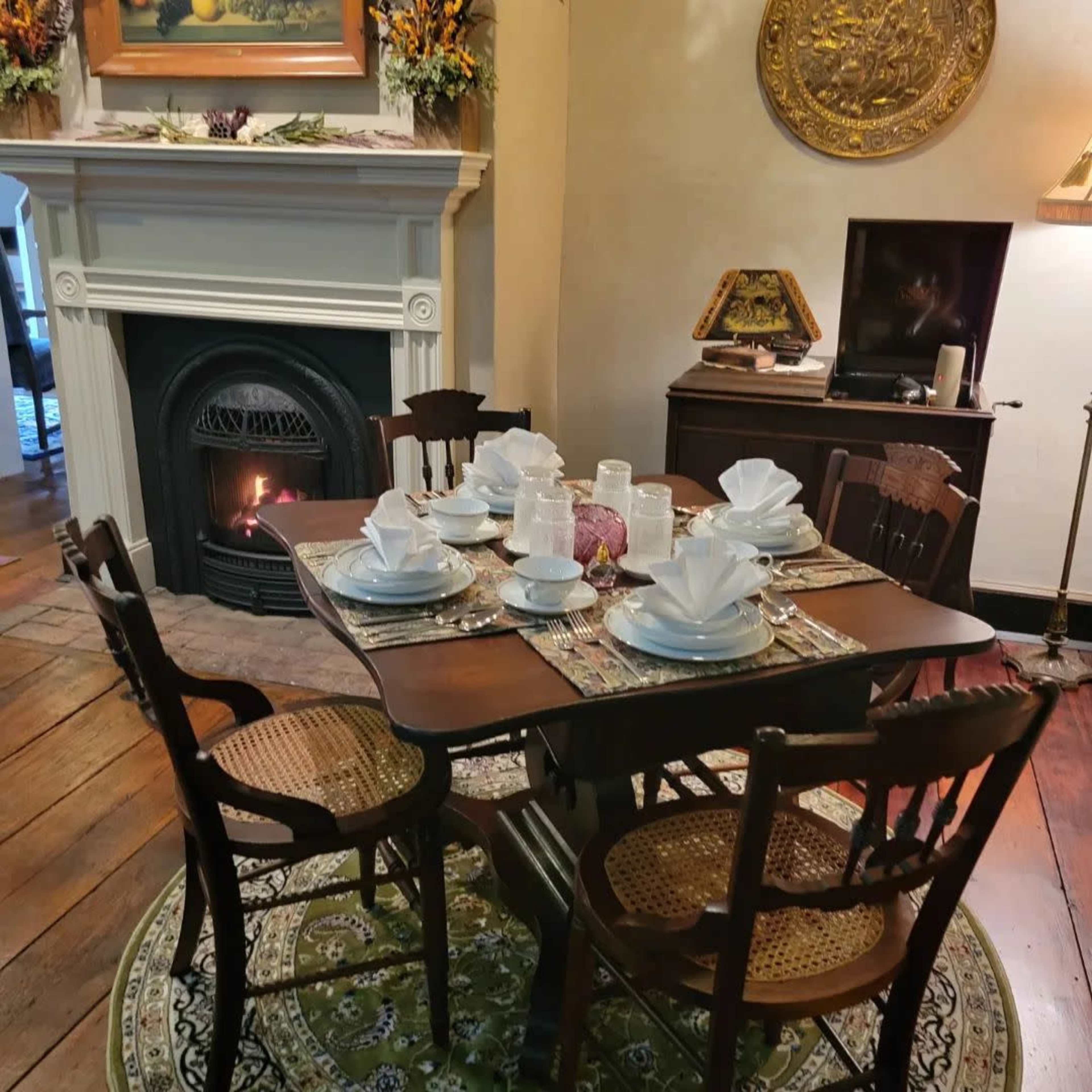 A dining table is set for a meal with neatly folded napkins, dishes, and glassware, surrounded by chairs in a cozy living space featuring a fireplace and decorative items.