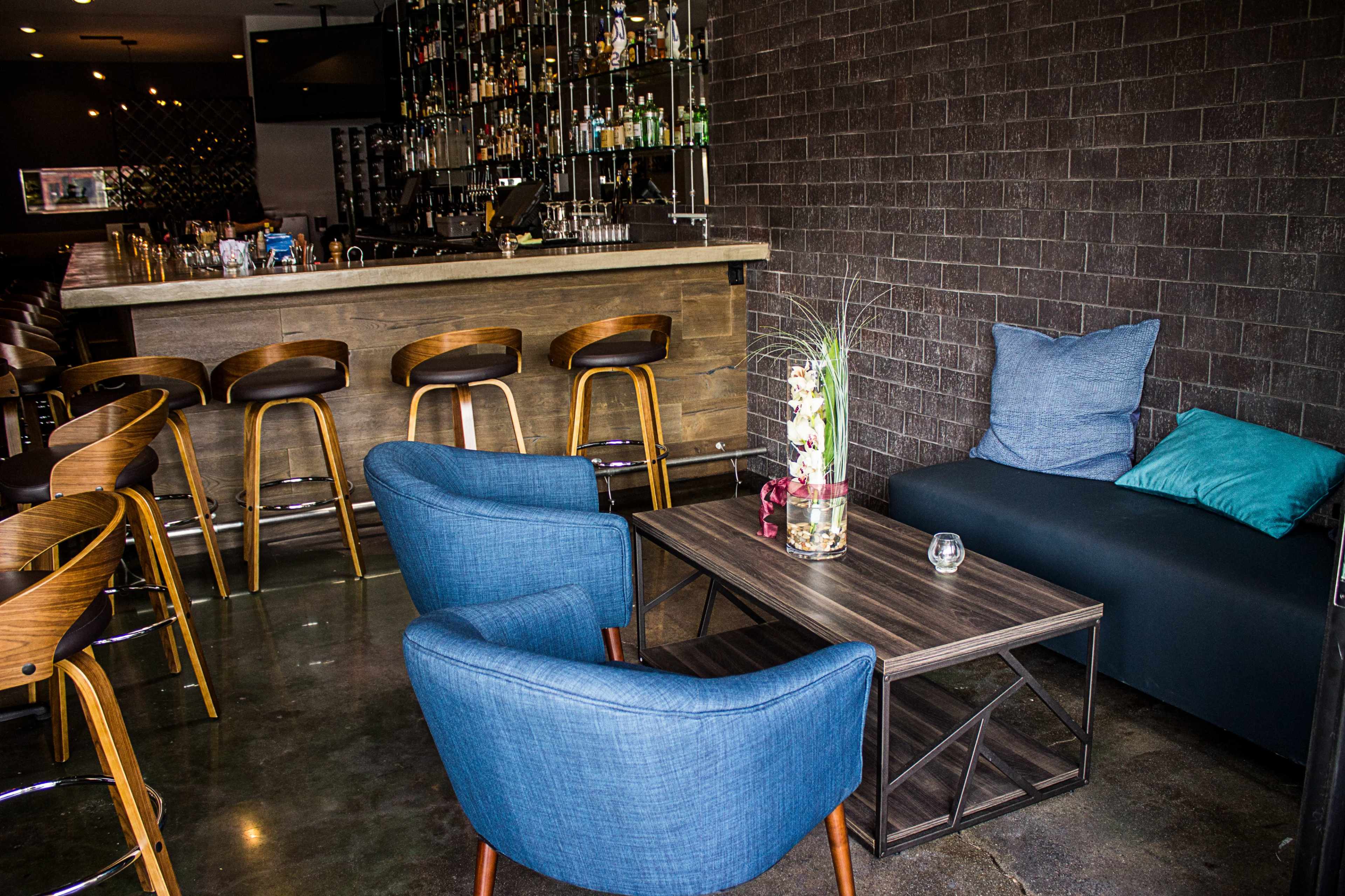 A modern lounge area featuring two blue chairs and a dark table set against a bar with wooden stools and a wall of shelves filled with bottles.