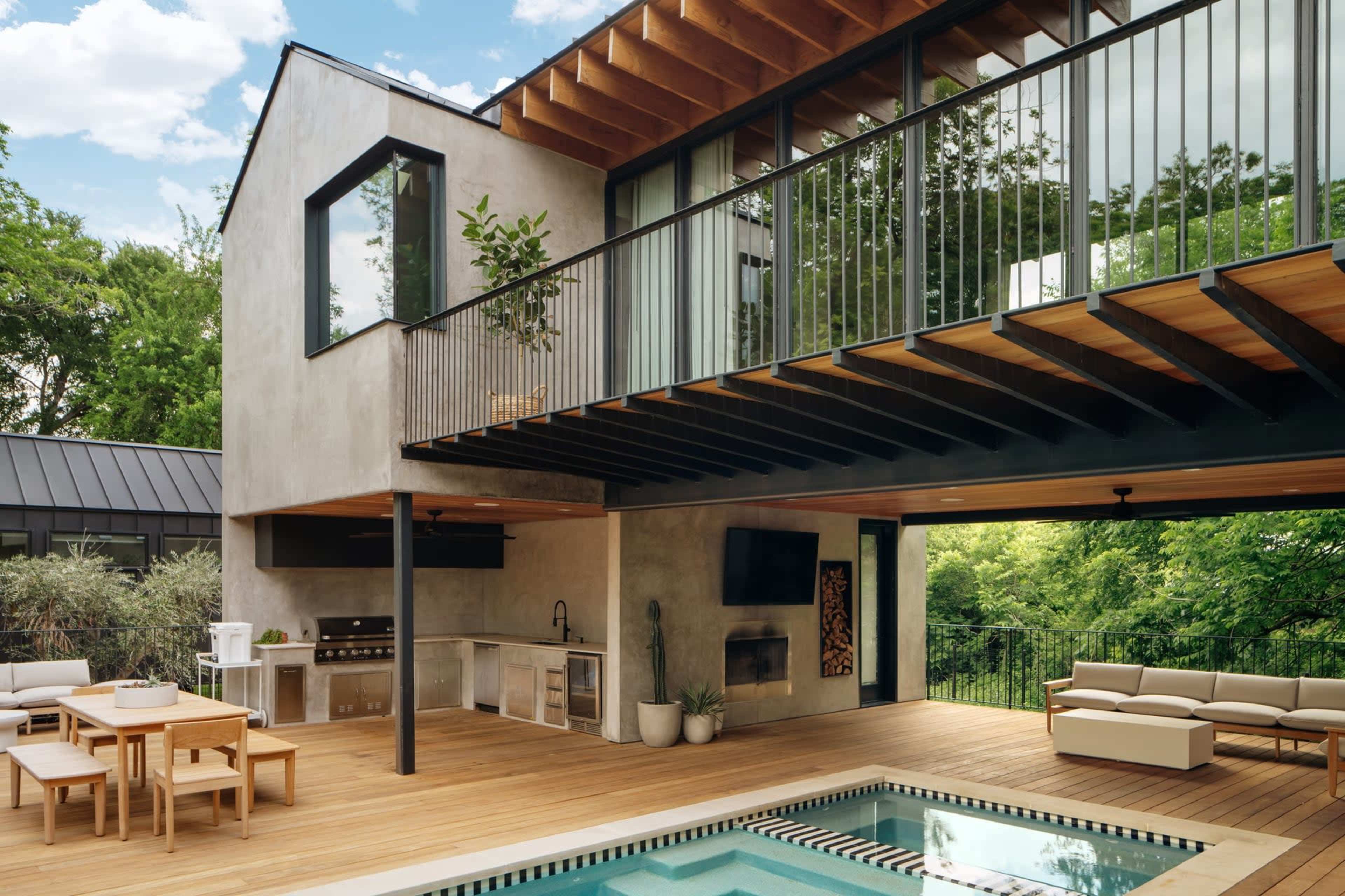 The image shows a modern house with a wooden deck featuring a pool, outdoor seating, and a grill area under a cantilevered upper level with large glass windows.