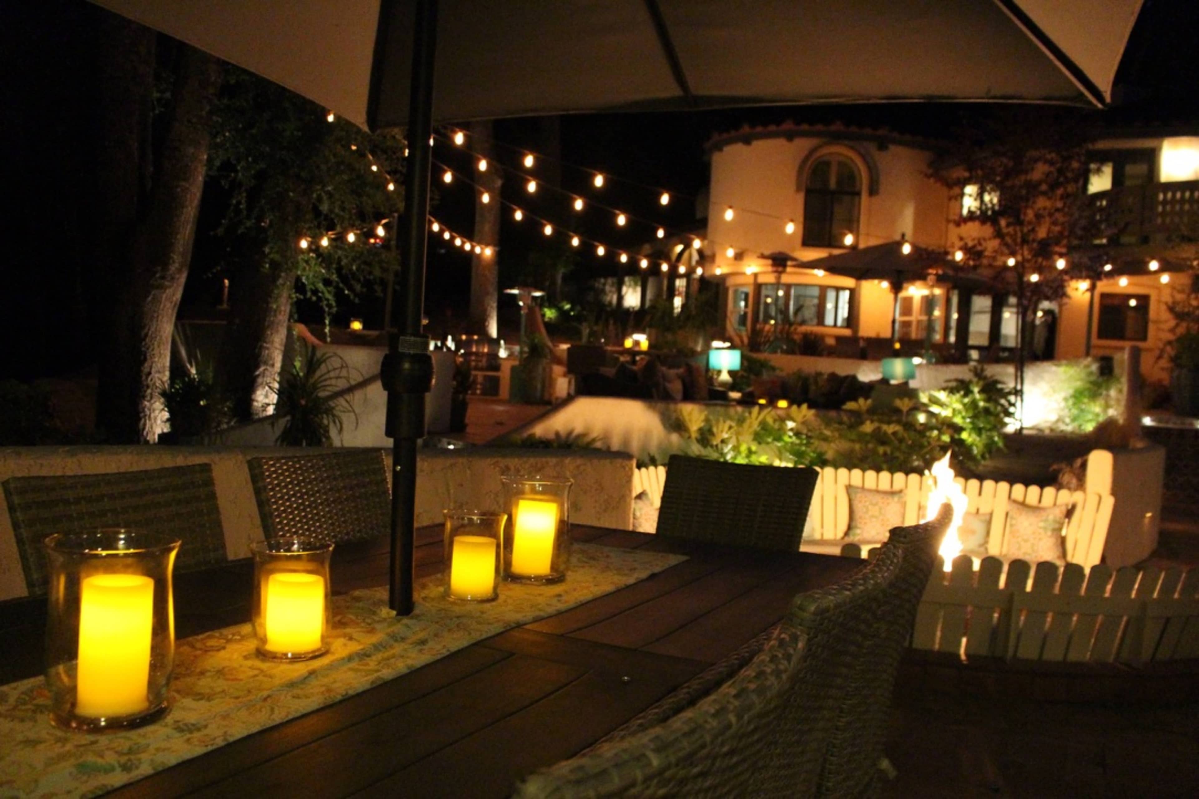 A well-lit outdoor patio features citronella candles on a table, string lights illuminate the surrounding area, and a house is visible in the background.