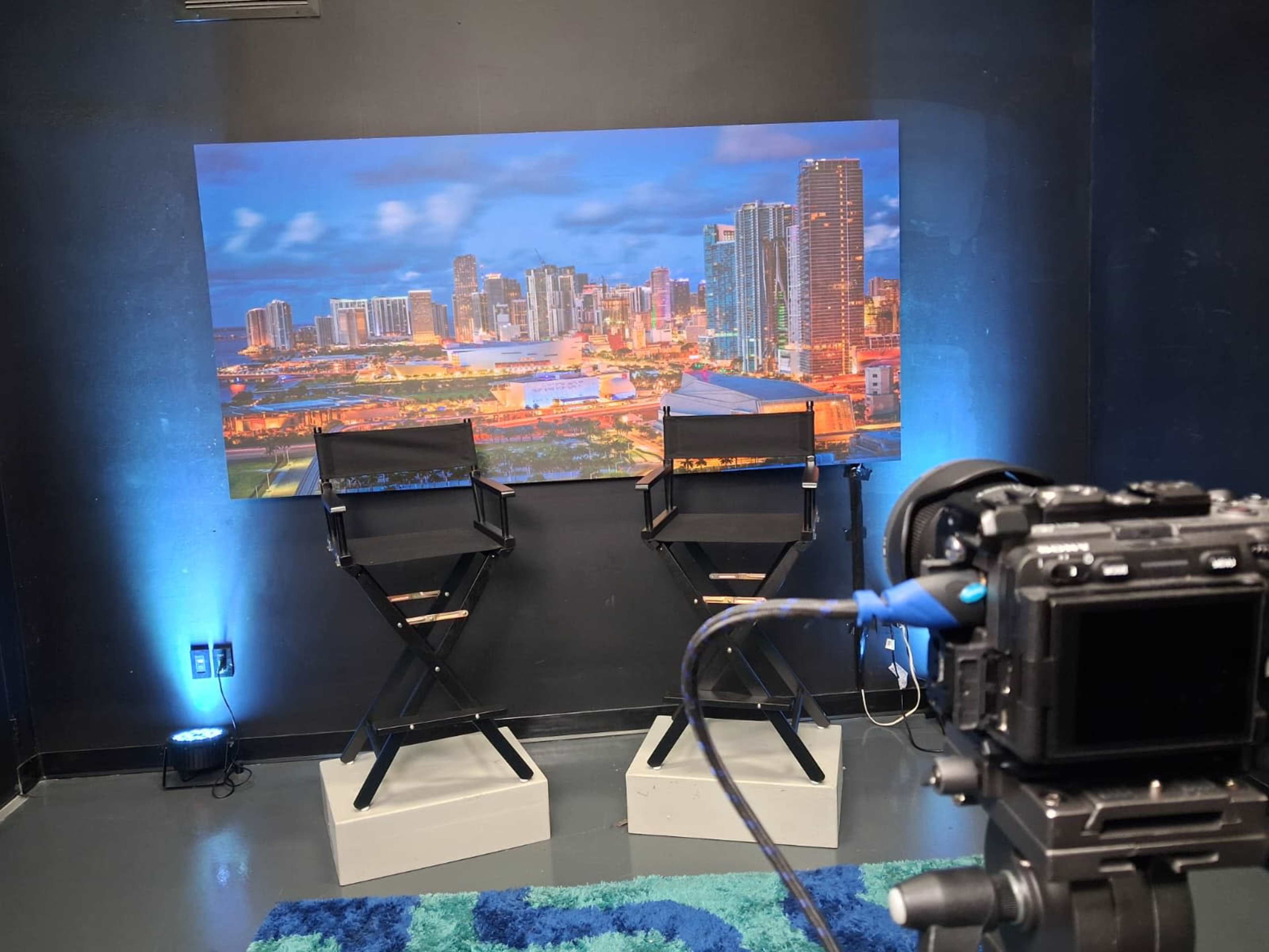 The image shows a studio setup with two director's chairs positioned in front of a backdrop of a city skyline at dusk and a camera on a tripod aimed at the chairs.