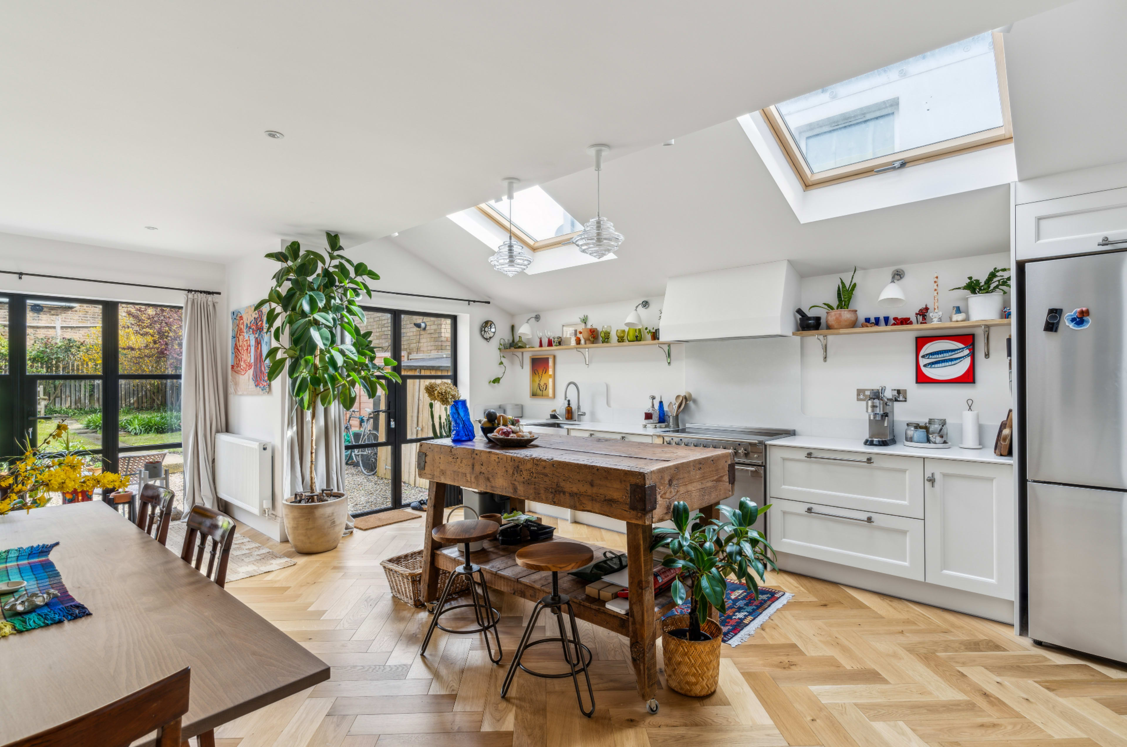A modern kitchen features a central wooden island with bar stools, large windows, and open shelving displaying plants and kitchenware.