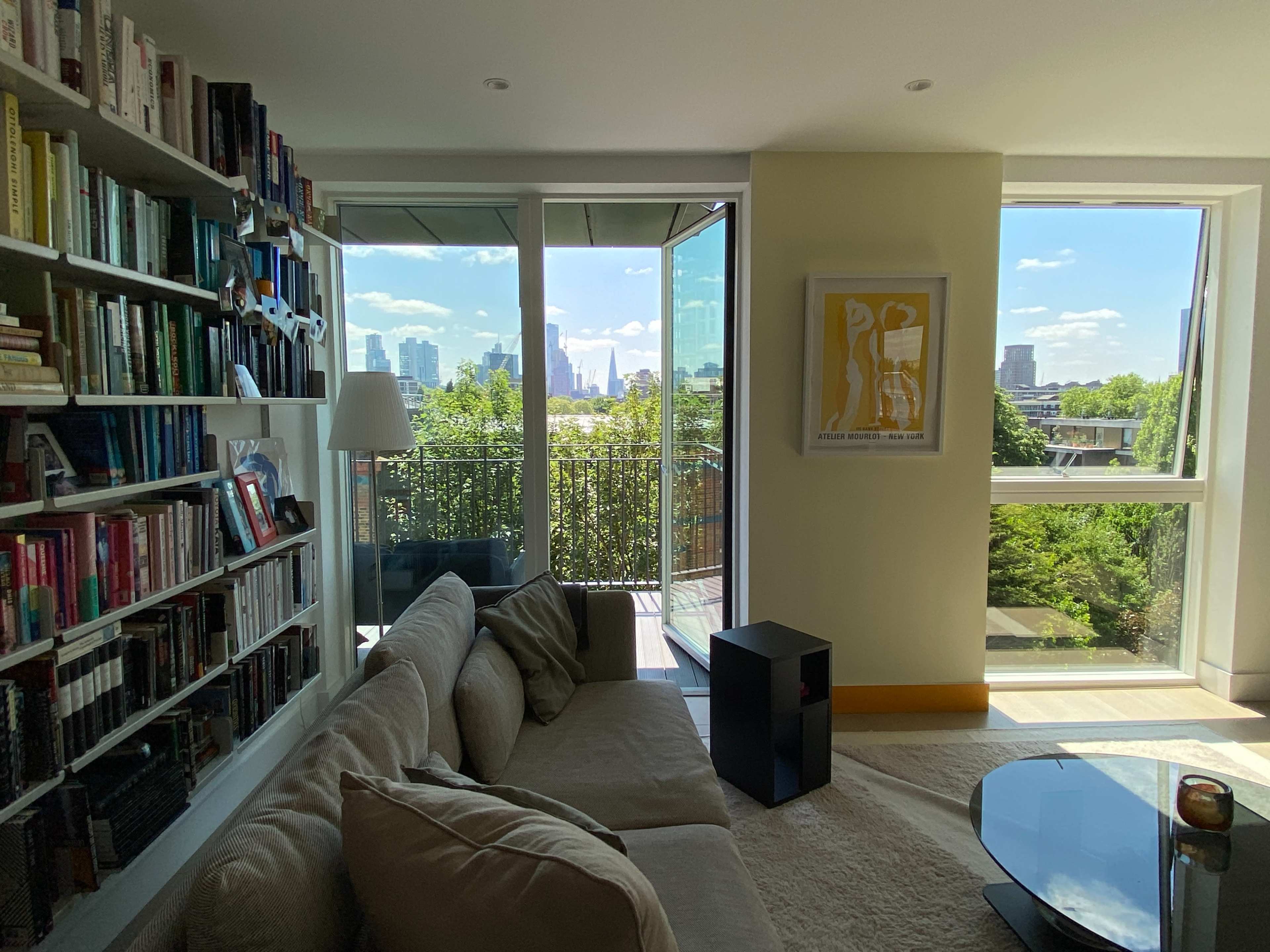 The image shows a modern living room featuring a beige couch, a small black table, a bookshelf filled with books, and large windows that provide a view of a city skyline and greenery outside.