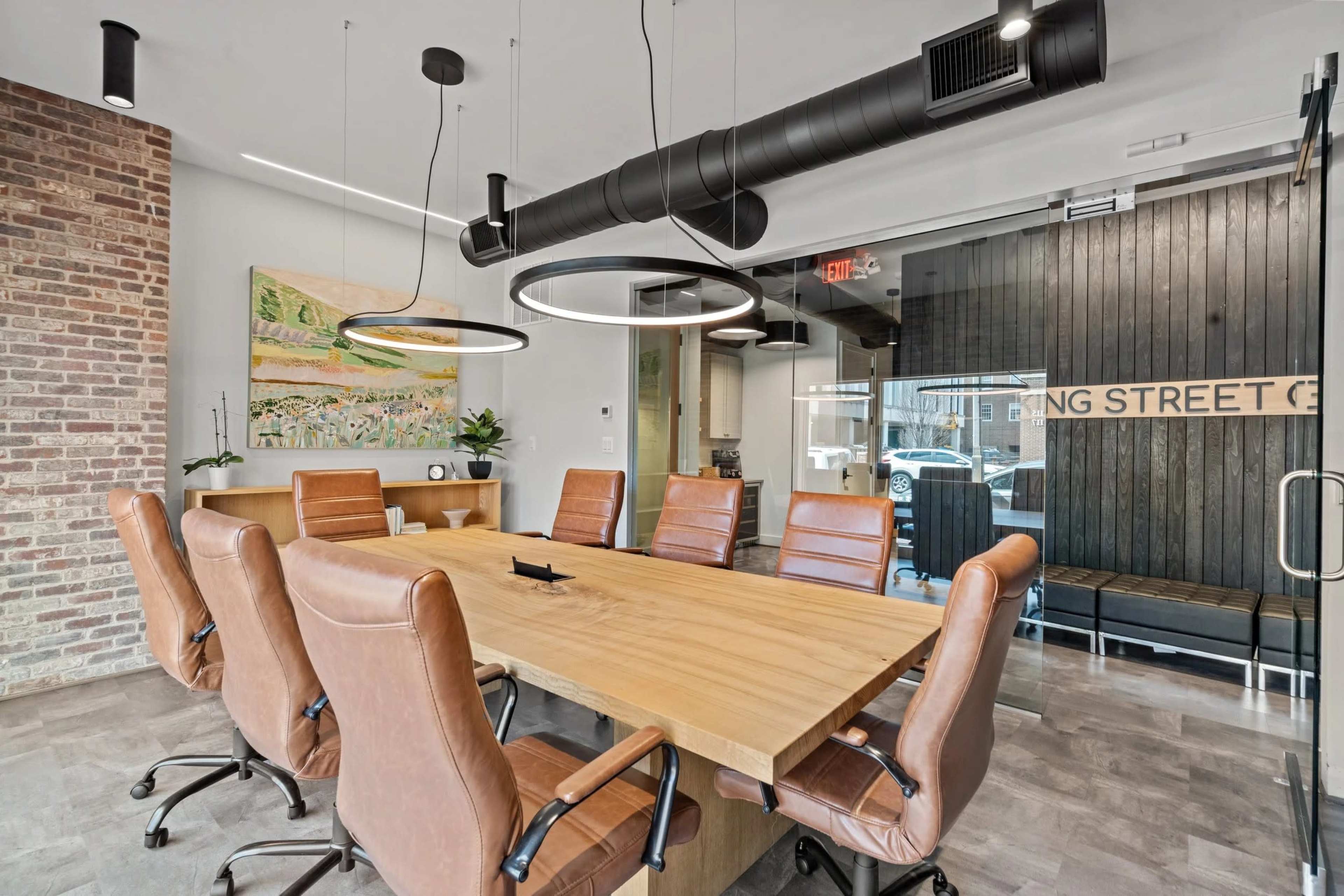 The image shows a modern conference room featuring a large wooden table surrounded by ergonomic leather chairs, with brick and glass walls.