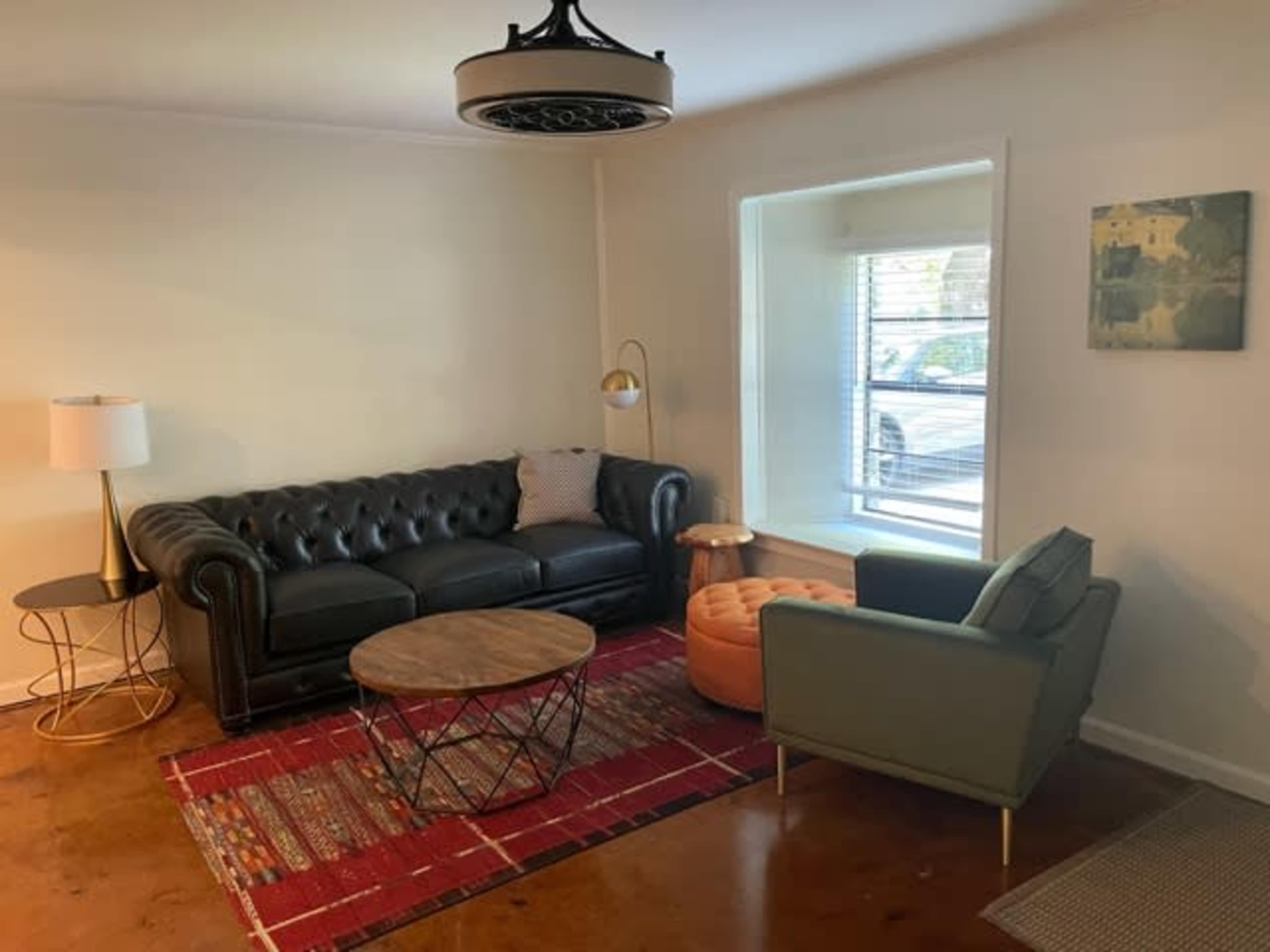 A black leather sofa and an olive green armchair are positioned around a circular coffee table on a patterned rug in a well-lit living room with a window.