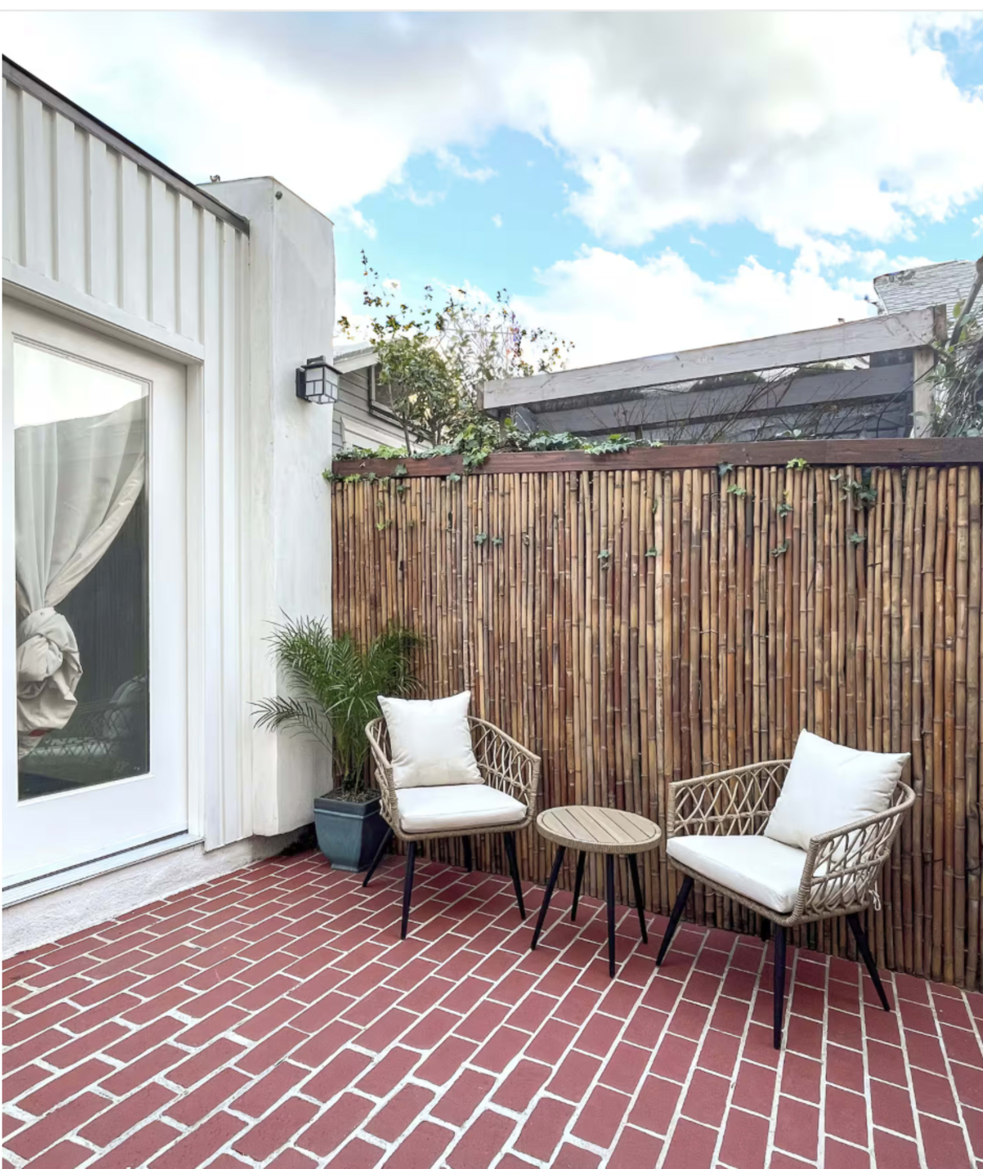 A small outdoor patio features two rattan chairs and a round table against a bamboo fence, with a potted plant nearby.