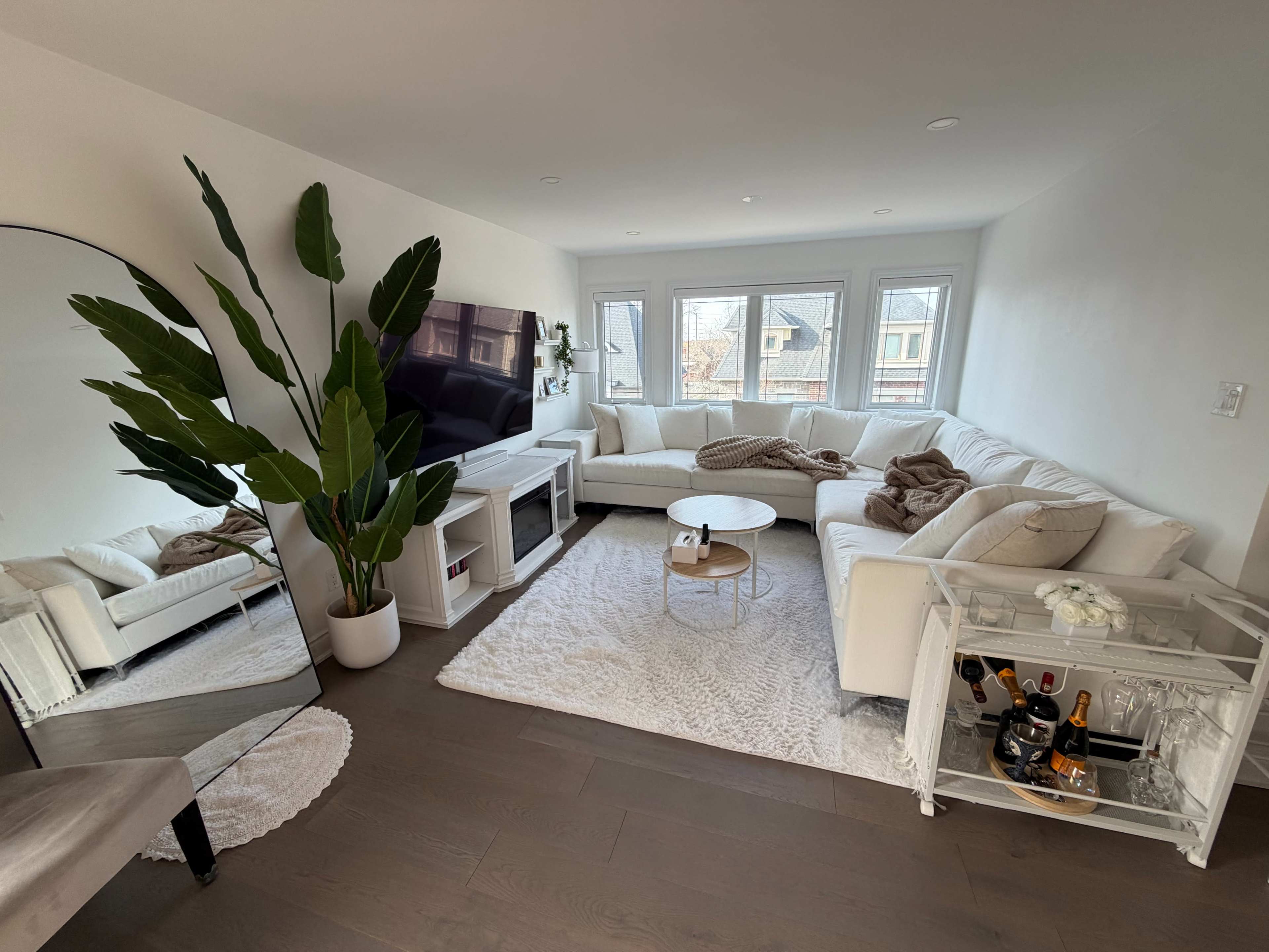 The image shows a modern living room featuring a white sectional sofa, a round coffee table, a large mirror, and a bar cart with drinks, all set against large windows.