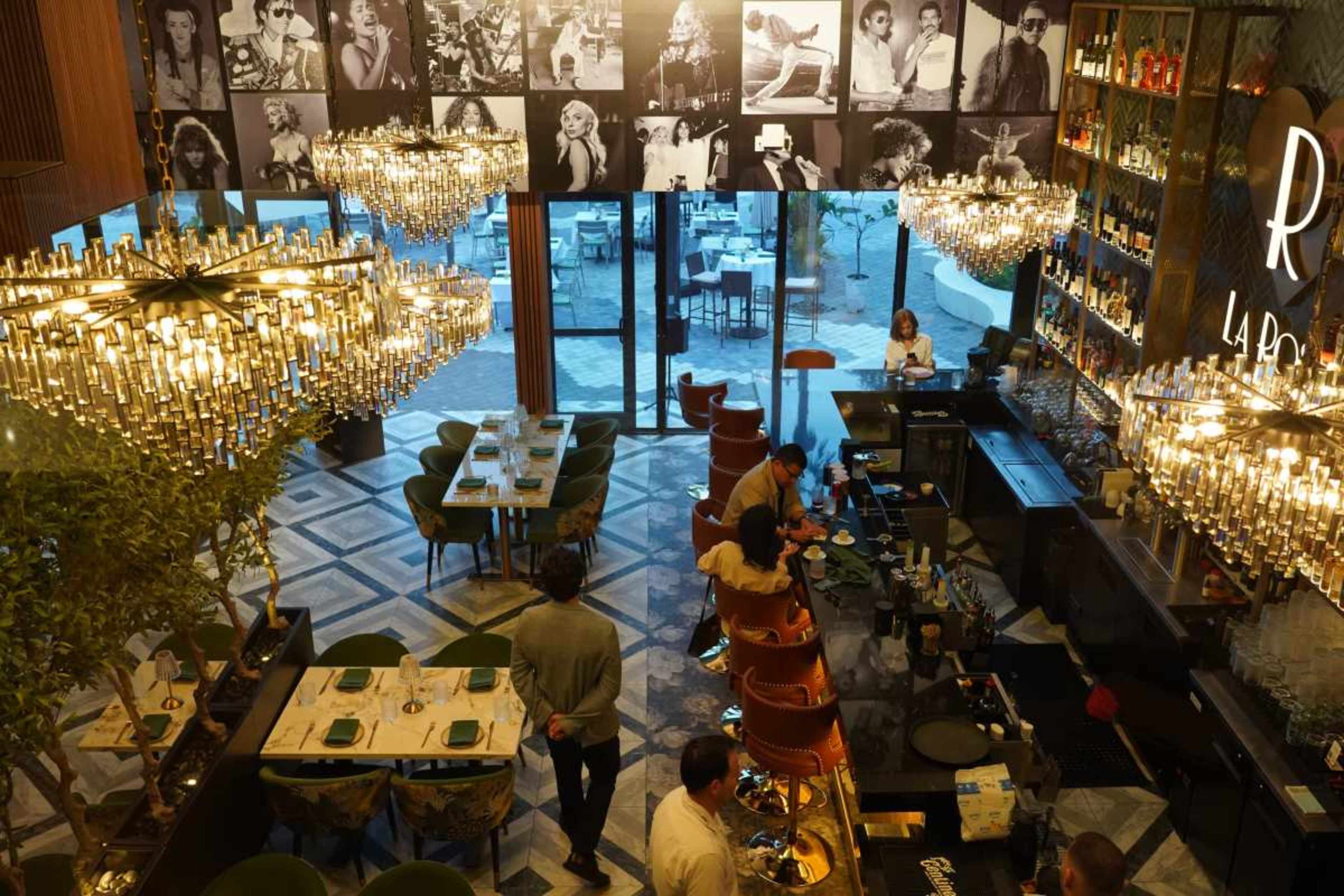 The image shows an upscale restaurant featuring chandeliers, a bar area, and walls adorned with black-and-white photographs of people.