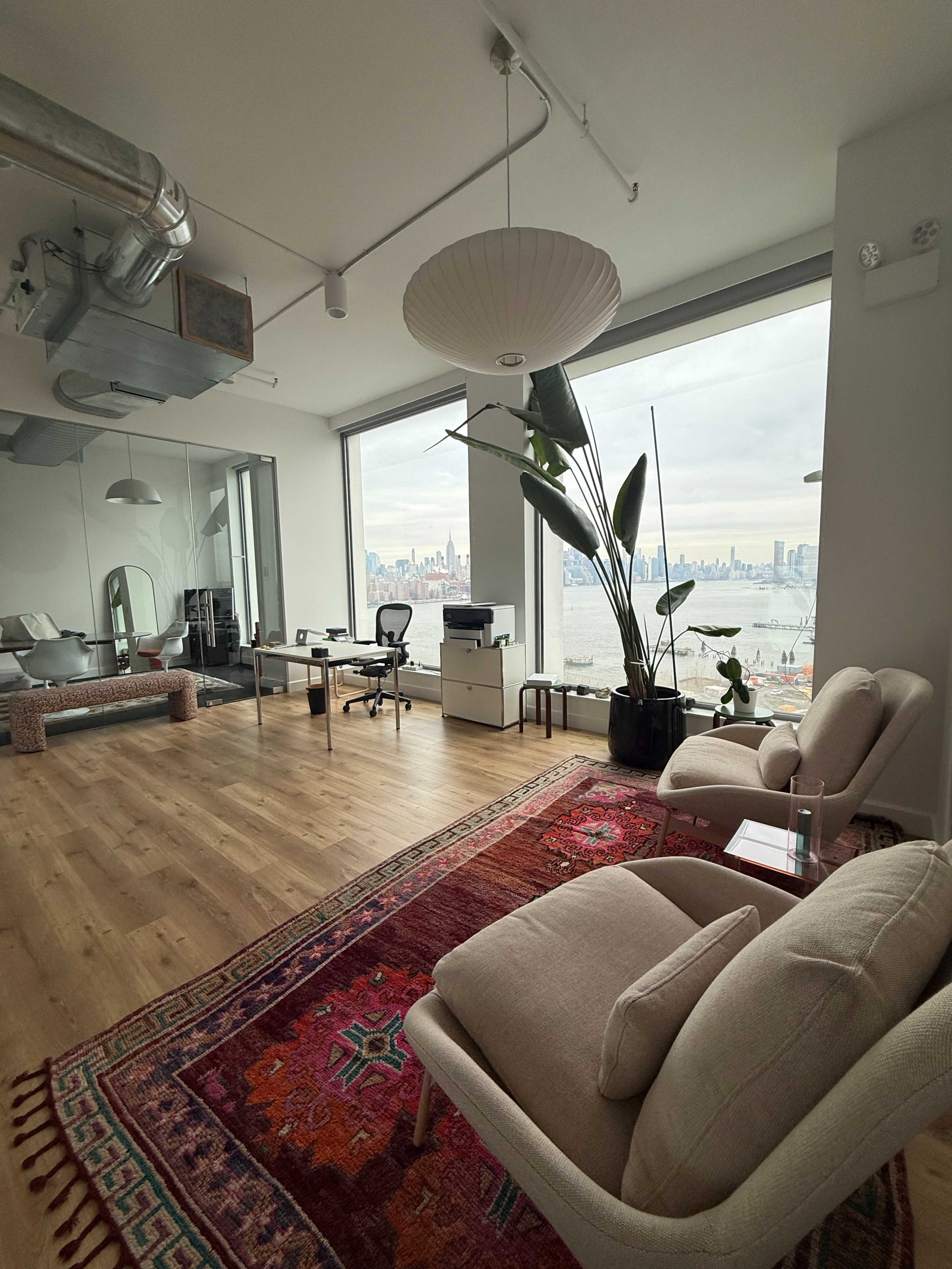 A modern office space features a seating area with two light-colored armchairs, a large window offering a view of the city skyline, and a workspace with a desk and chairs.