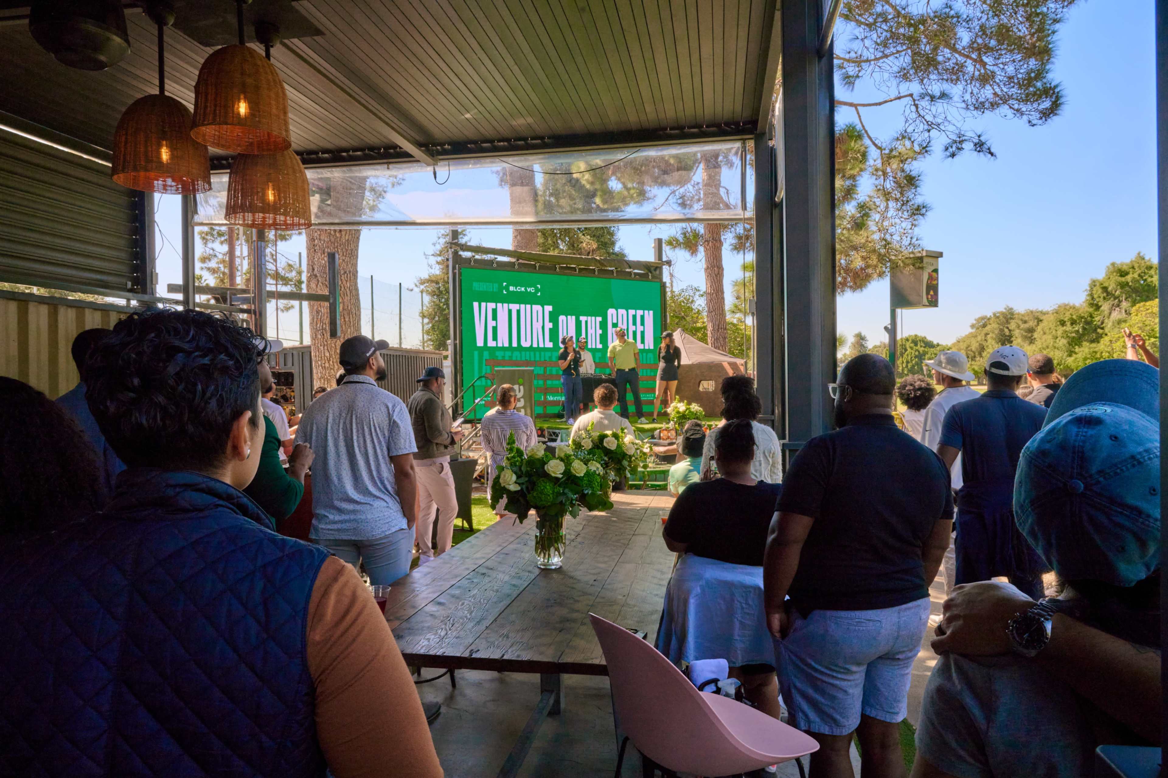 A crowd is gathered inside a venue with large windows, watching a speaker on a stage set against a green backdrop that says "VENTURE OUT THE PEER."