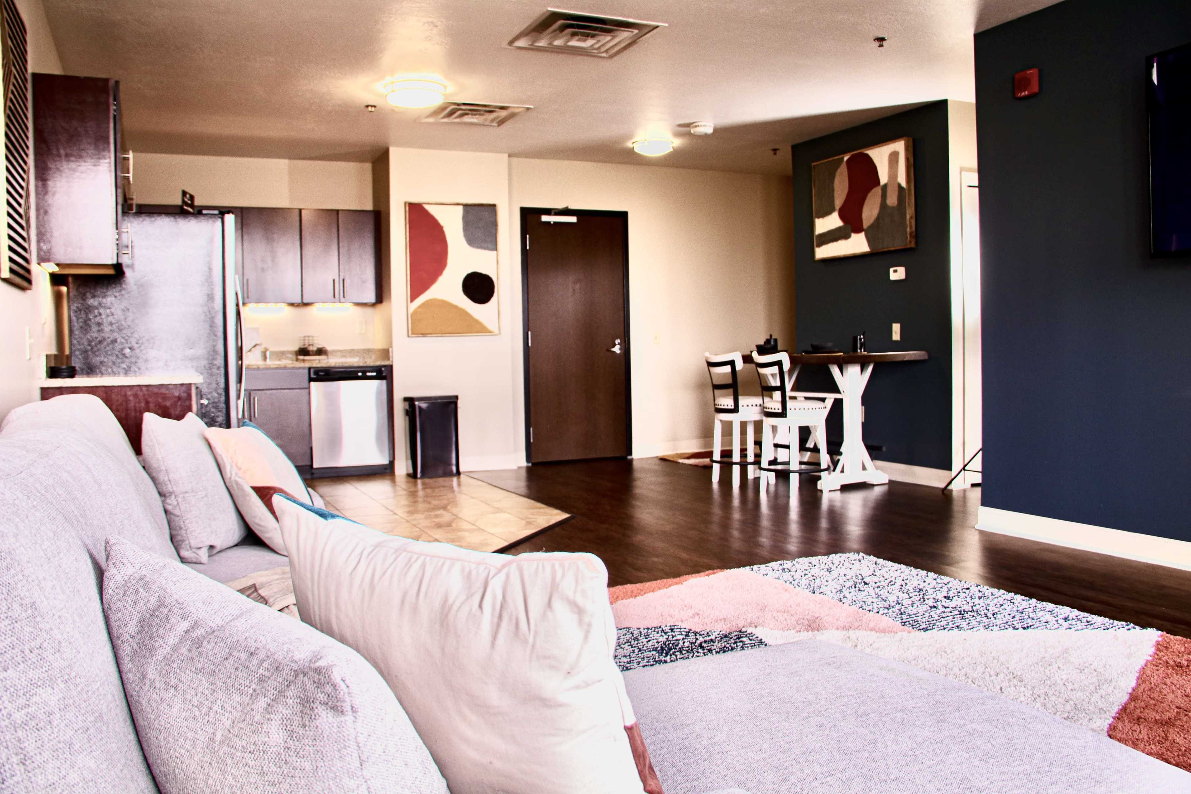 The image shows a modern living space with a couch in the foreground, a kitchen area in the back, a dining table to the right, and abstract artwork on the walls.