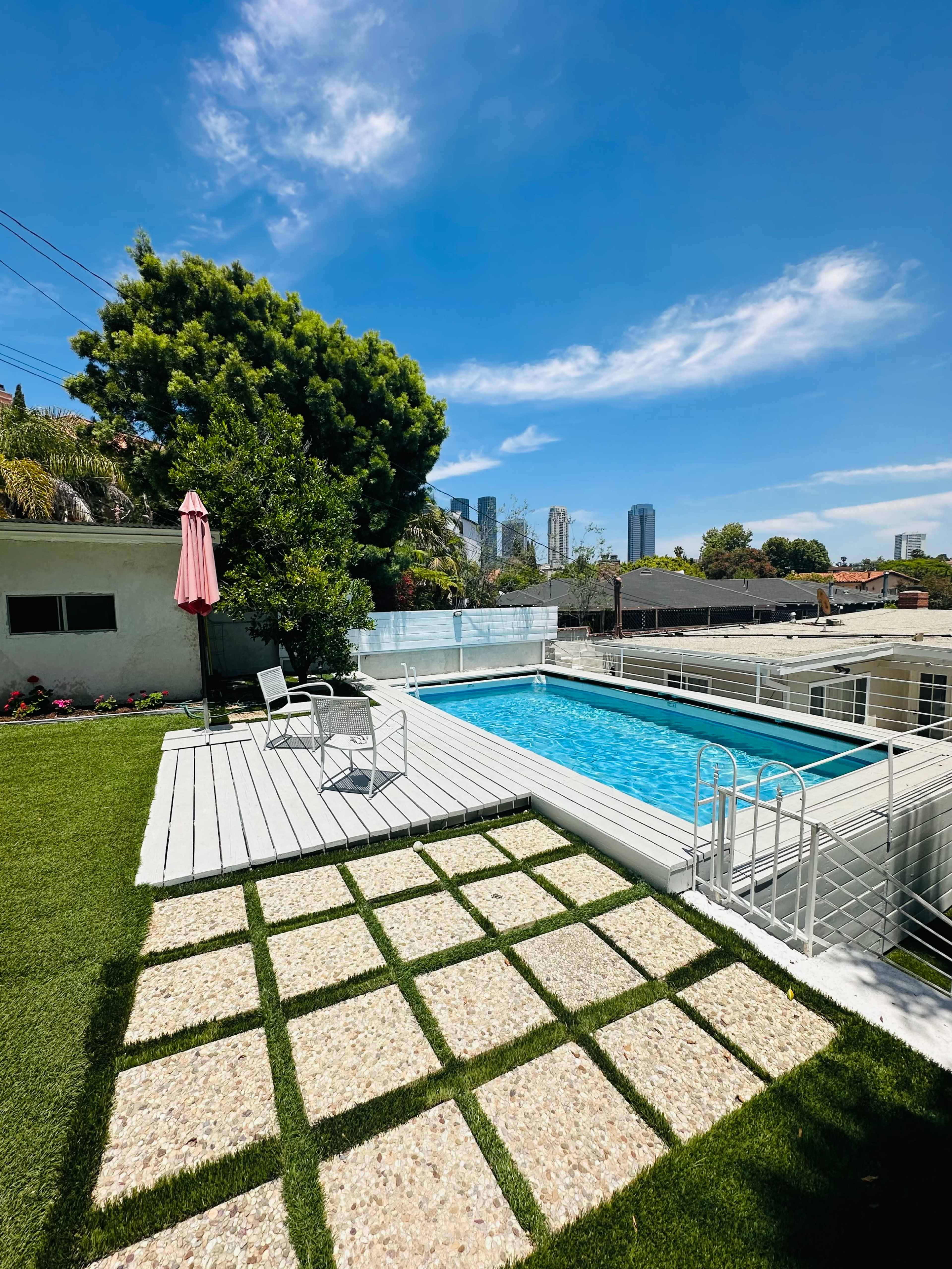 A modern backyard with a swimming pool, surrounded by green grass and stone pathways, and city buildings visible in the background.