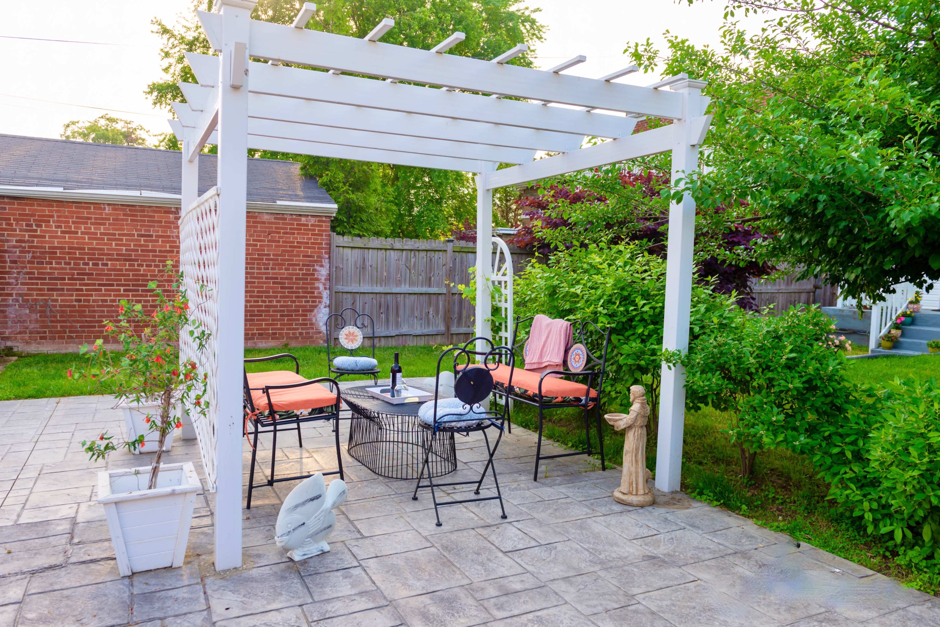 A white pergola with seating and a small table is set up on a stone patio surrounded by greenery.