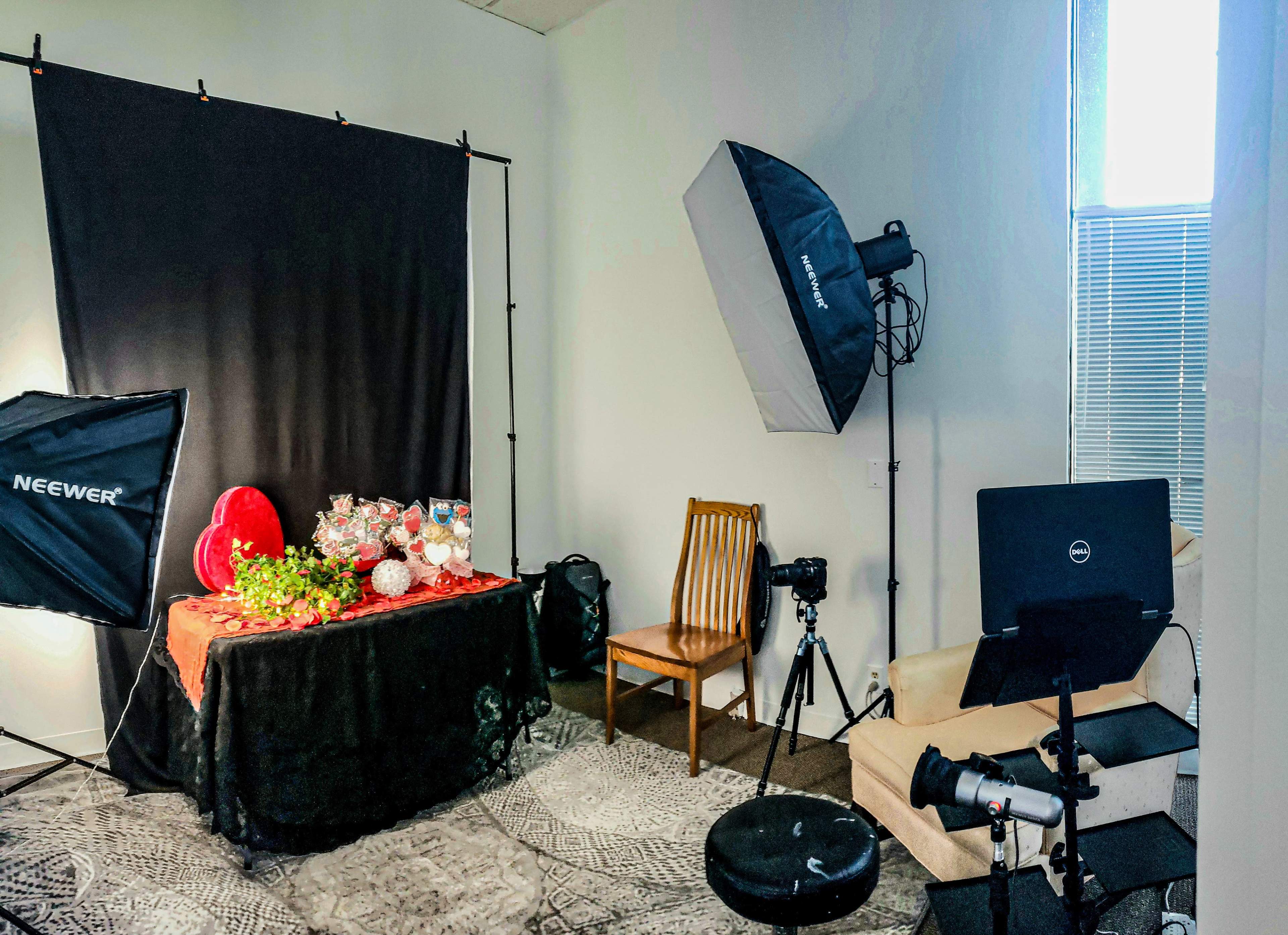 The image shows a photography setup in a room featuring a black backdrop, lighting equipment, a table with decorative items, and a camera on a tripod.