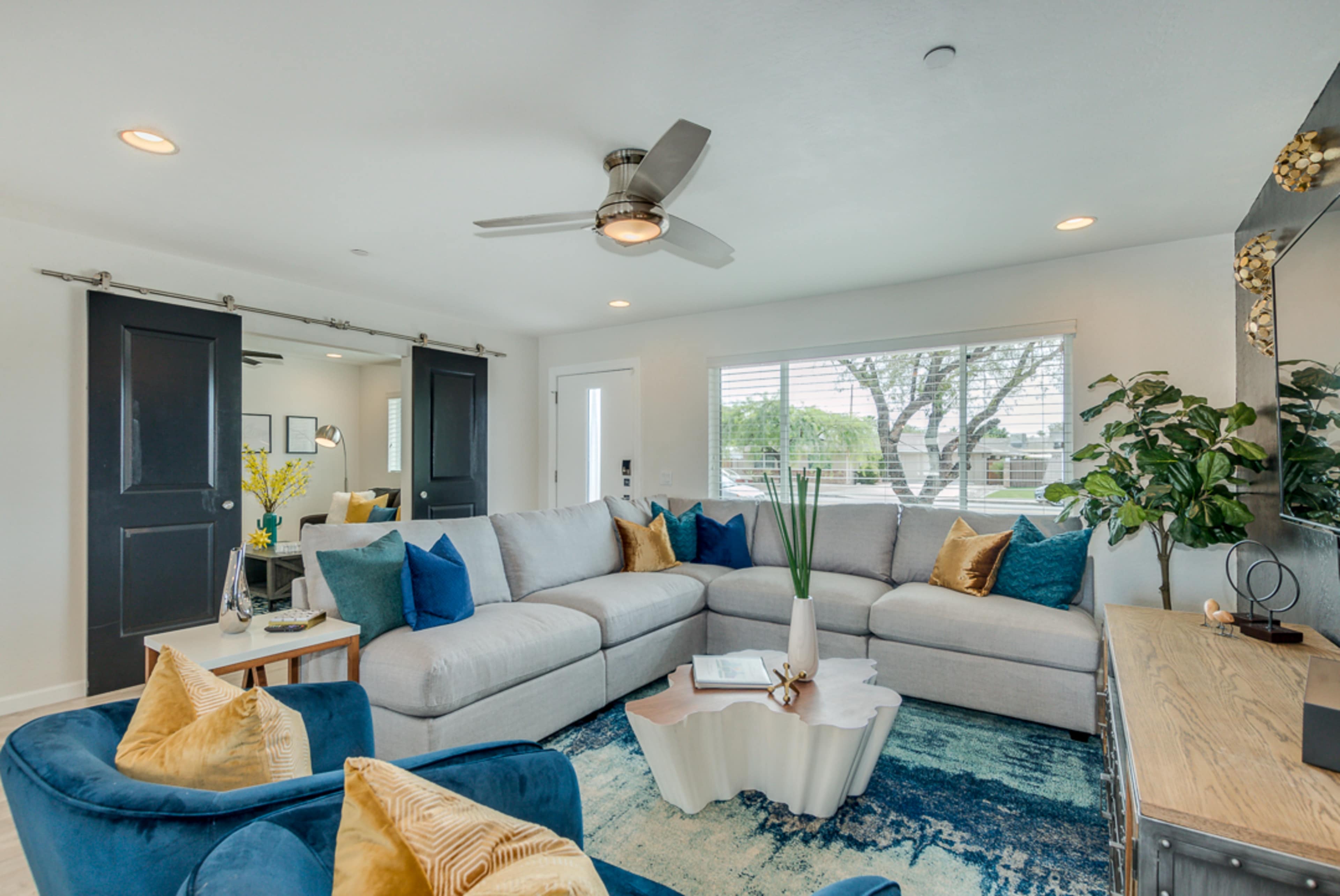 The living room features a large gray sectional sofa, blue accent chairs, and a decorative coffee table arranged around a rug, with light streaming in through a large window.