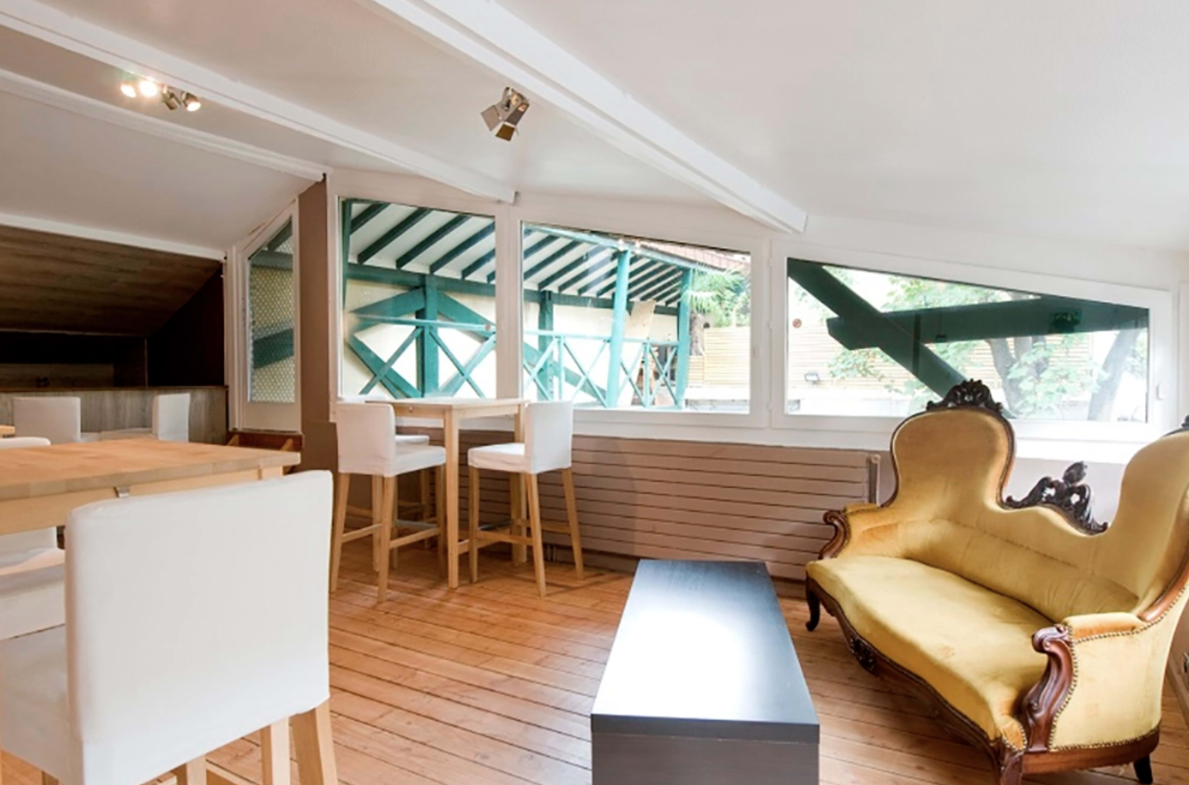 A bright, open interior space featuring a yellow upholstered sofa, a wooden dining table and chairs, and large windows with green trim.