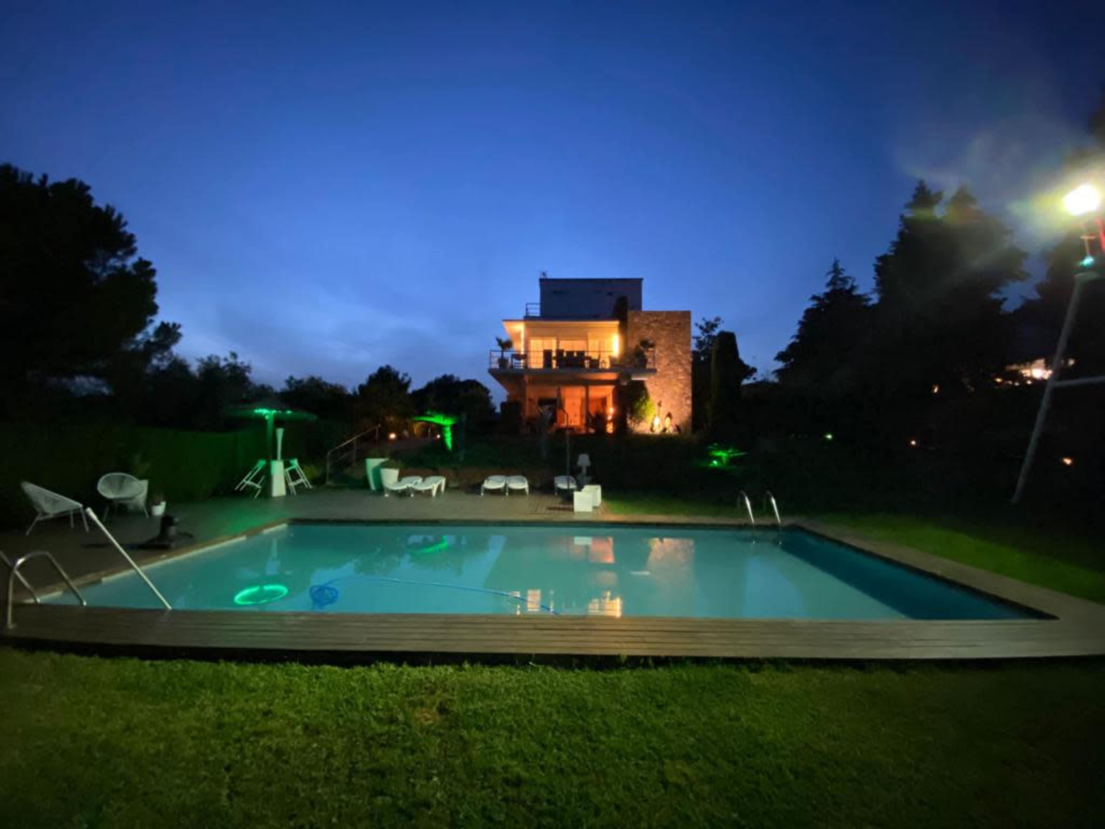 A modern house with stone accents stands behind a well-lit swimming pool surrounded by lush greenery at night.