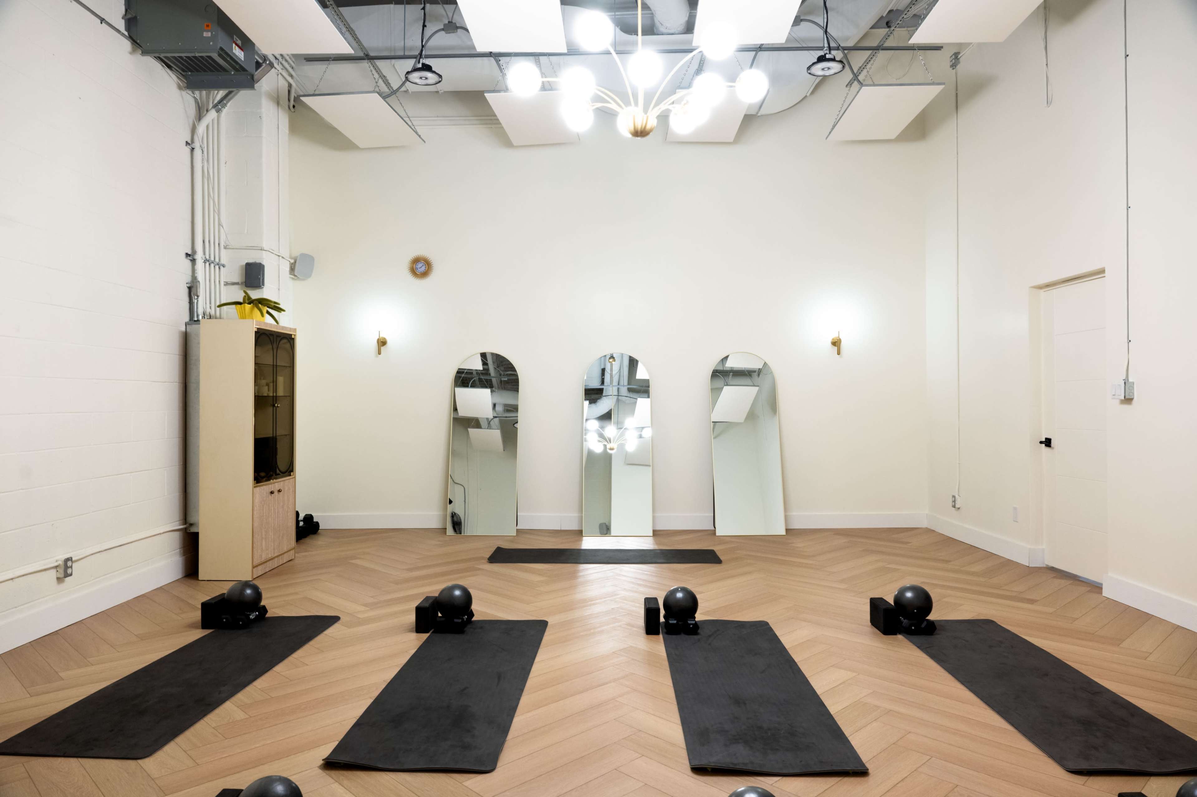 The image shows a yoga or exercise studio with several workout mats, dumbbells, and three mirrors reflecting the room's lighting fixtures.