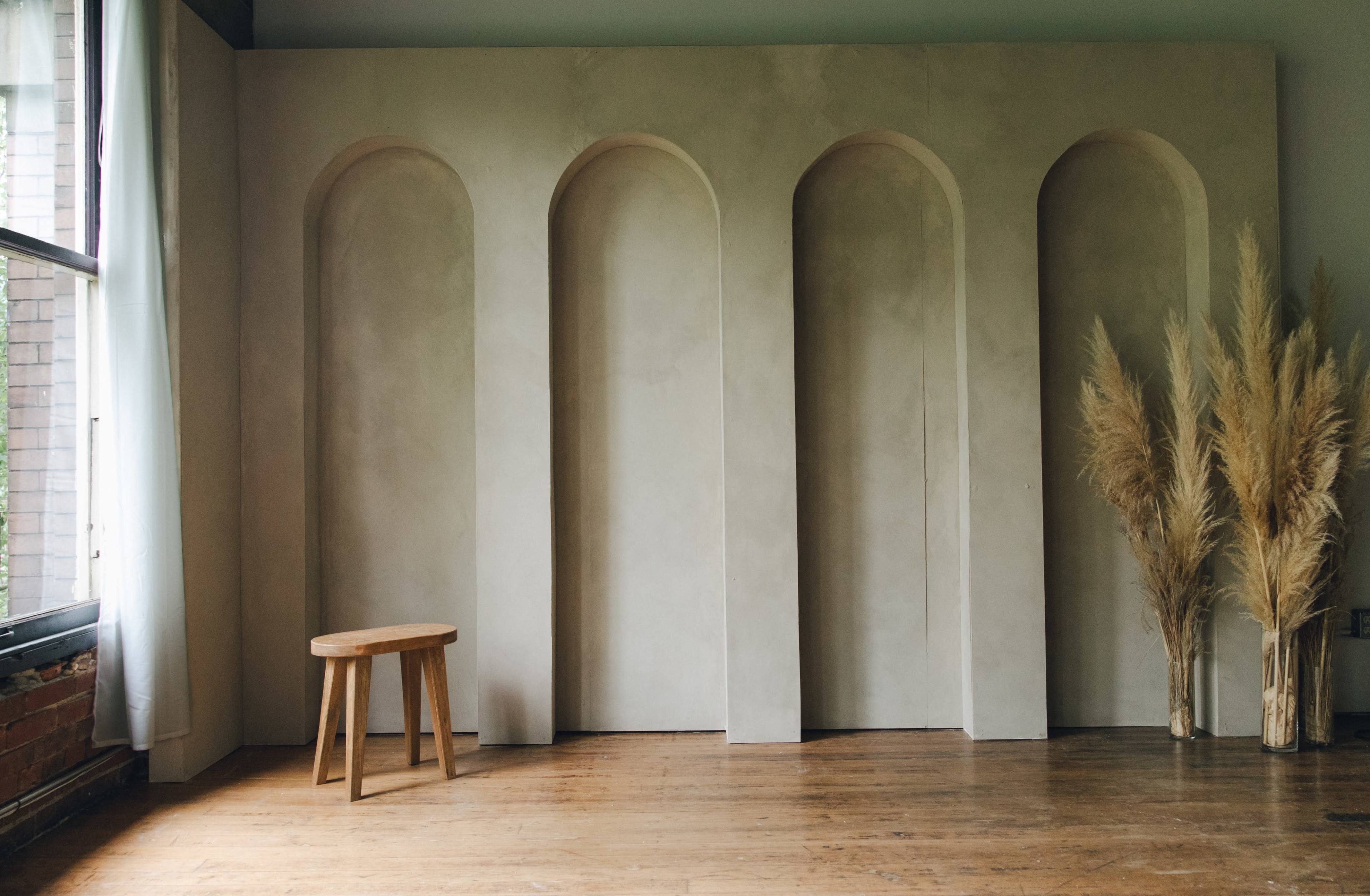 A textured wall with three arched niches and a wooden stool in front of it, alongside a cluster of dried pampas grass.