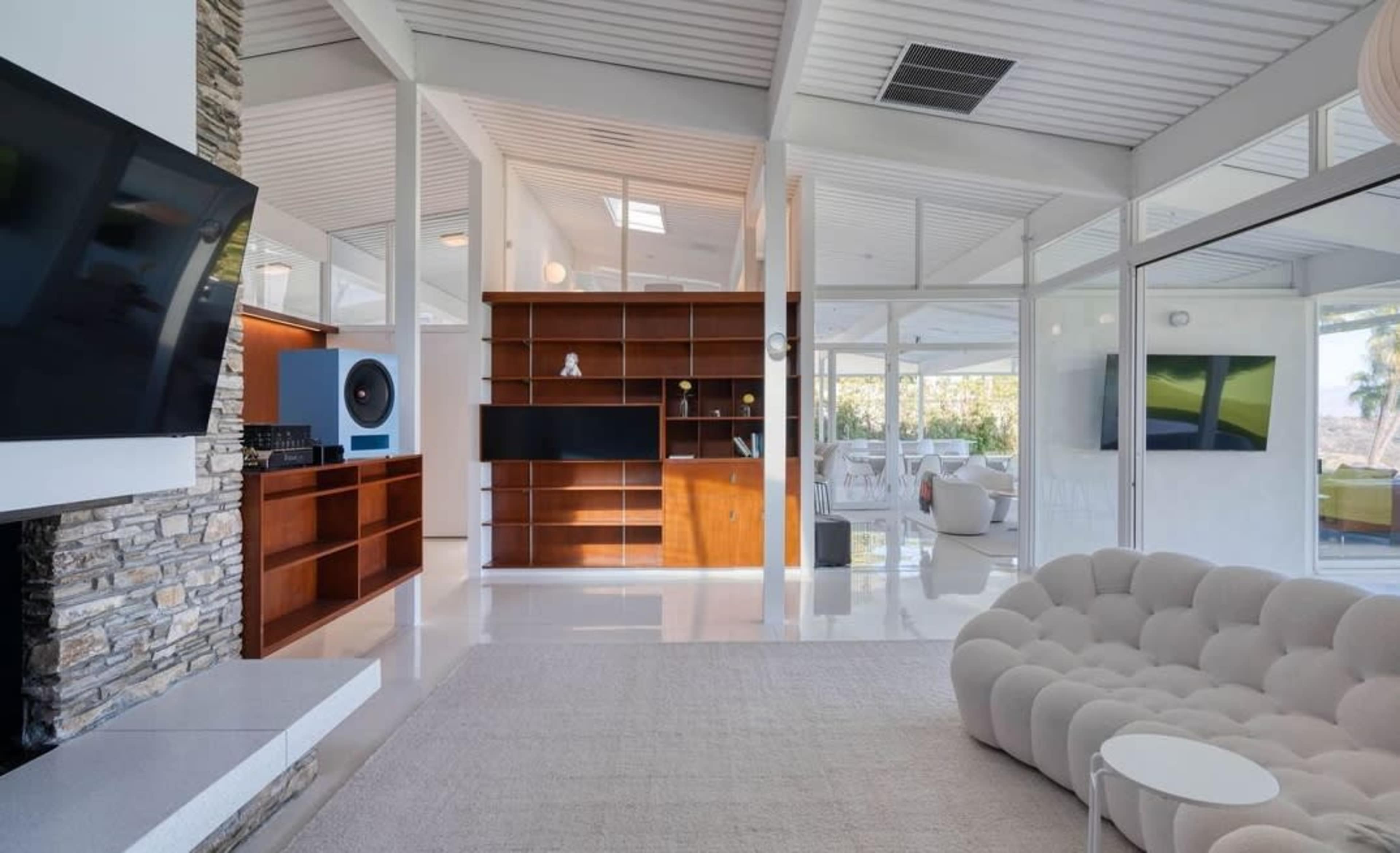The image shows a modern, open-concept living space featuring a white stone fireplace, a large curved sofa, and a wooden shelving unit.