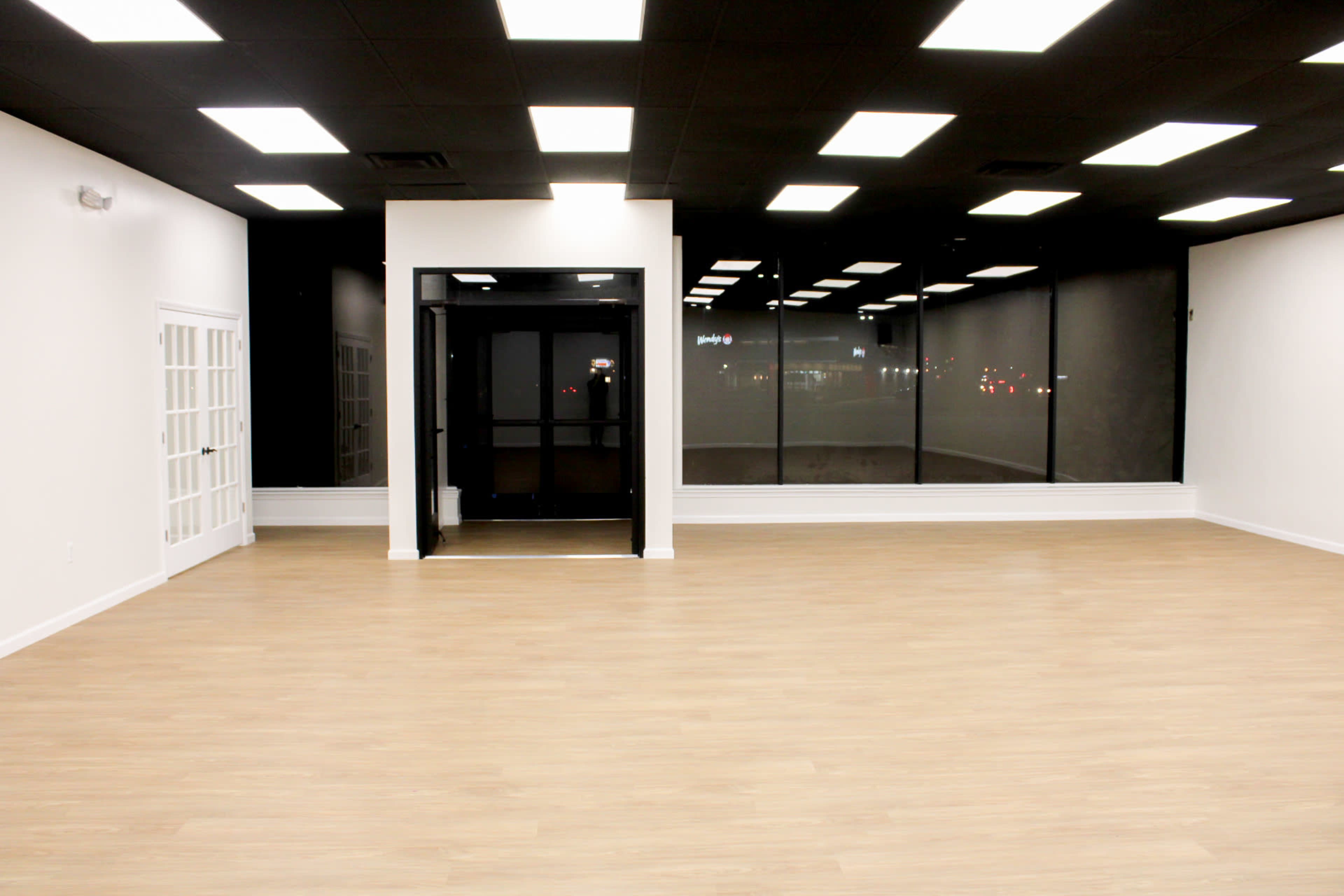 The image shows an empty retail space with a large window facing the street, light-colored walls, and a wooden floor.