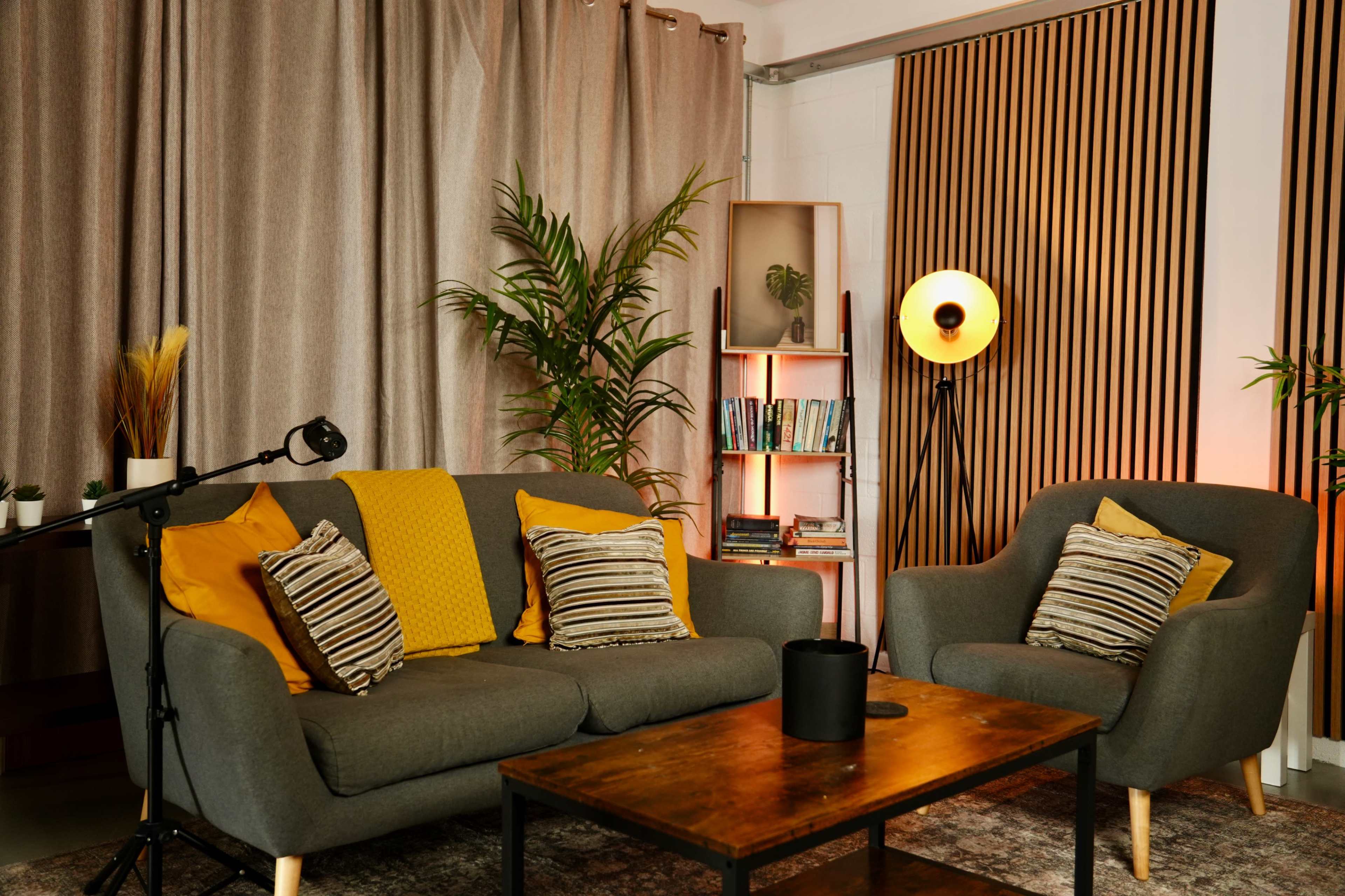 The image shows a modern living room with a gray sofa and armchair, complemented by yellow pillows, a wooden coffee table, plants, and a bookshelf.