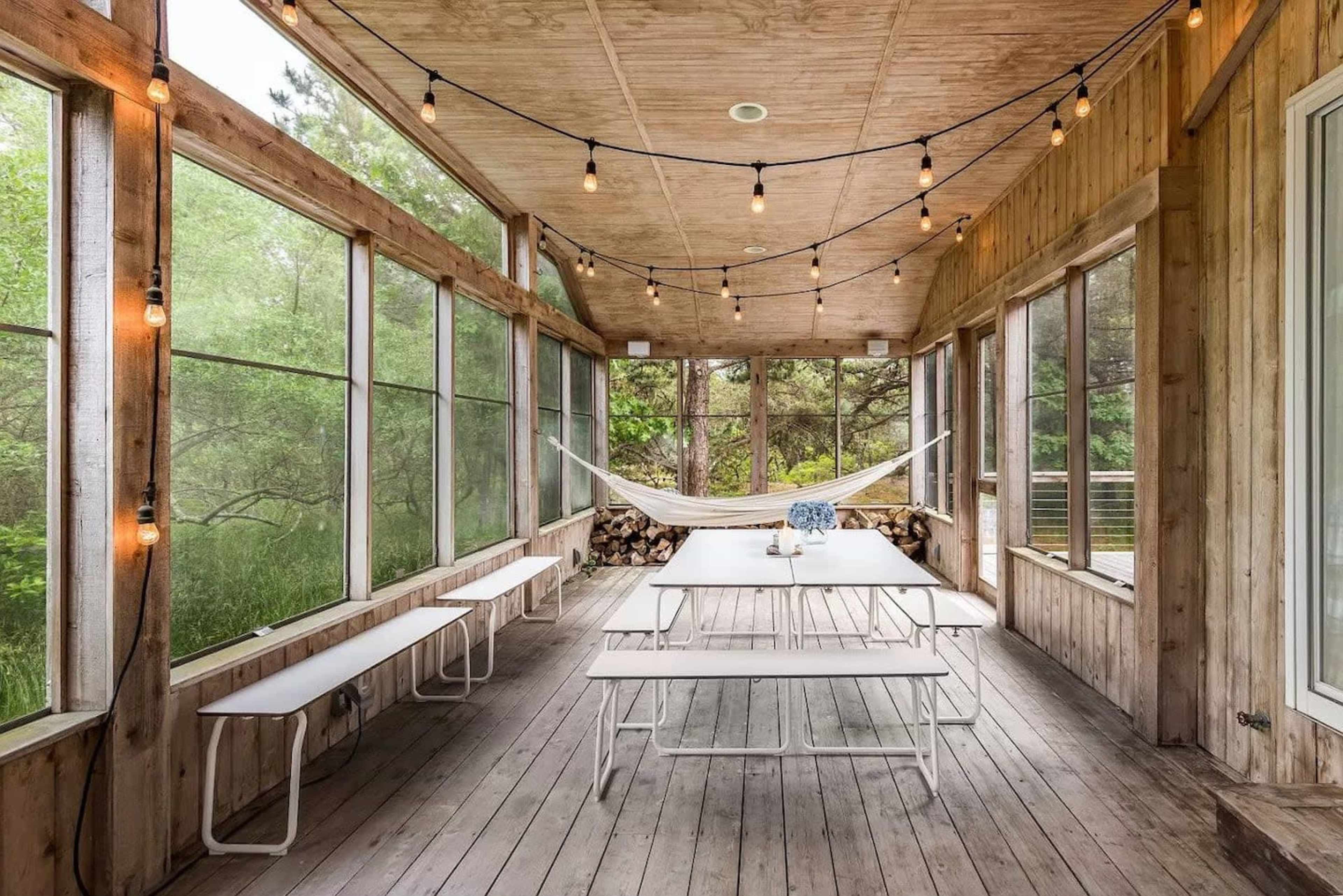 A porch with wooden walls and ceiling, featuring string lights, a long white table with benches, and a hammock in a natural setting.