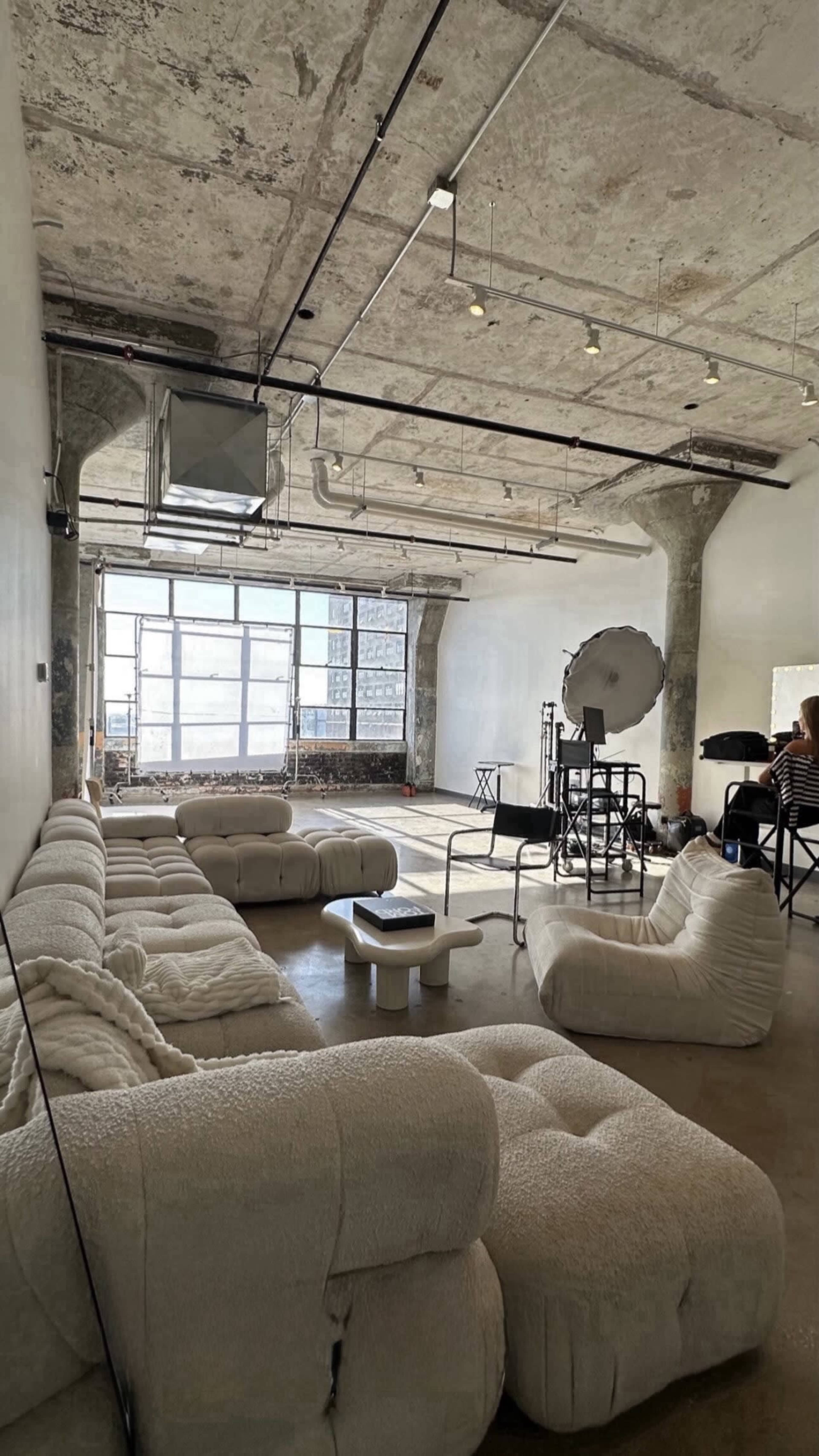 The image shows a spacious room with exposed concrete ceilings and large windows, featuring soft, white furniture arranged in a casual layout.