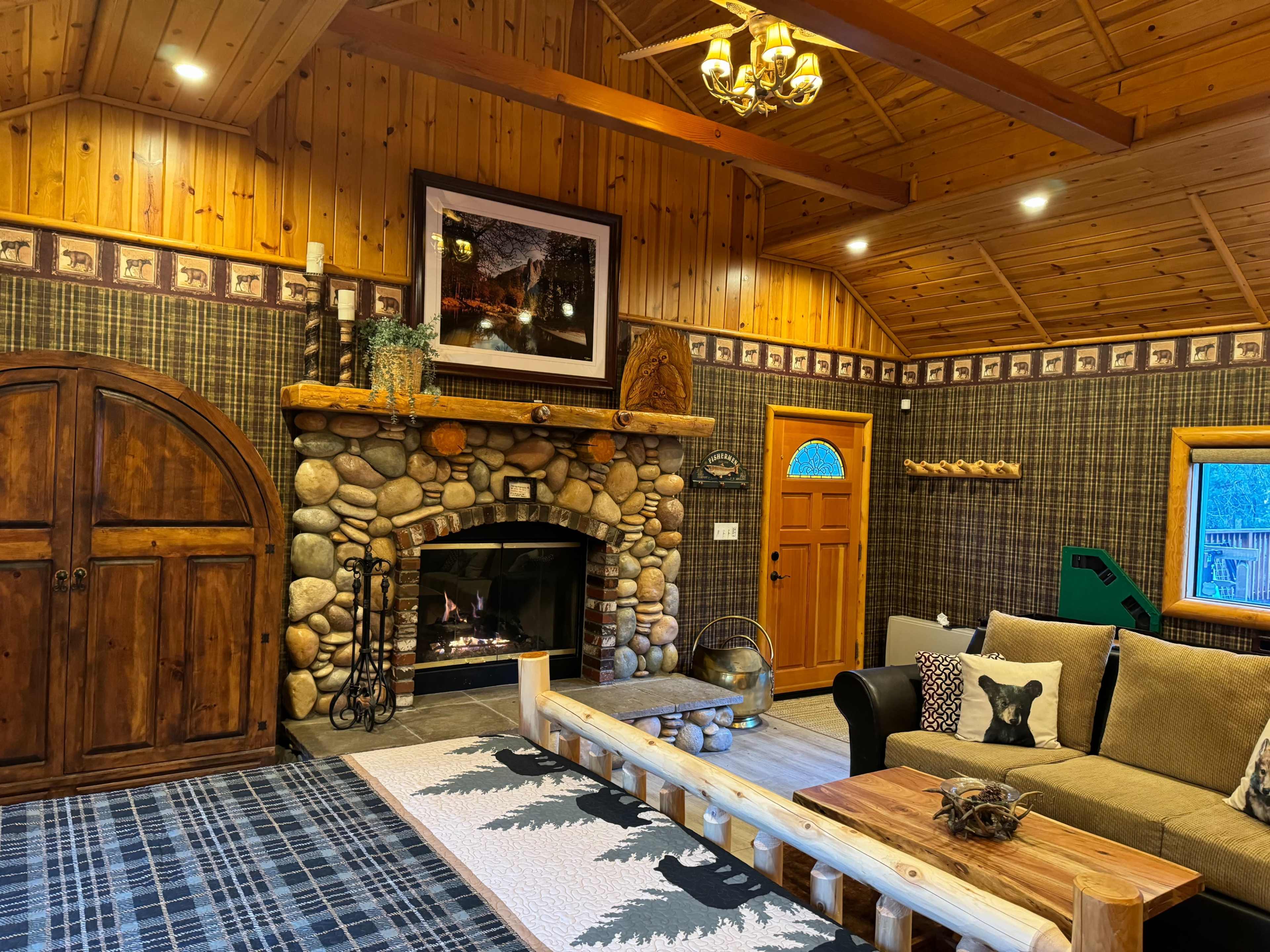 The image shows a cozy cabin living room featuring a stone fireplace, wooden walls, and a rustic seating area.