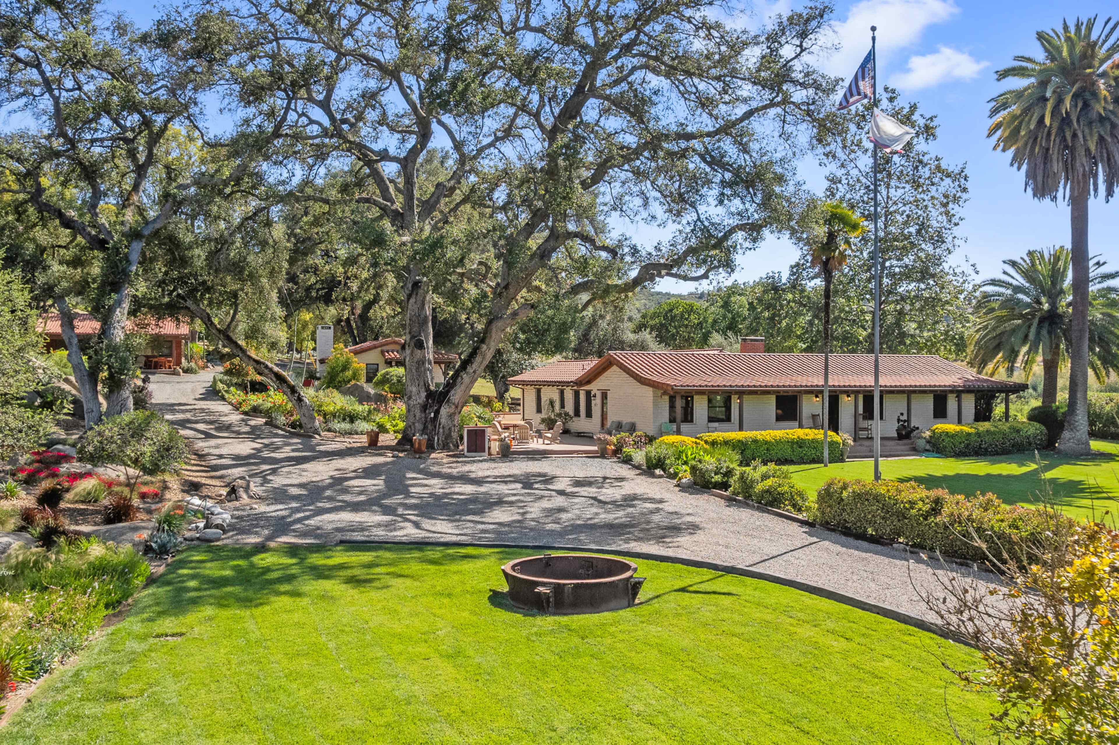 A sprawling ranch-style house with a red tile roof is surrounded by lush greenery and palm trees, featuring a circular stone fire pit in the foreground.