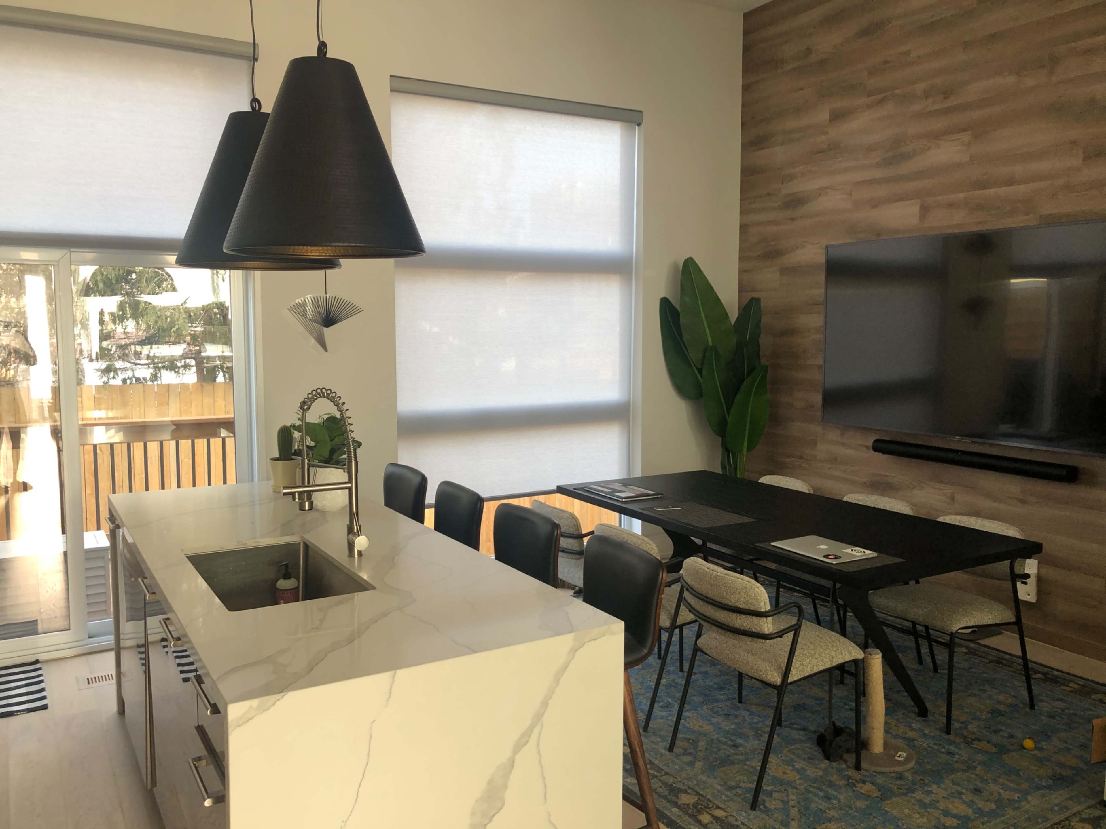The image shows a modern kitchen and dining area featuring a marble countertop, large black pendant lights, a dining table with chairs, and a wall-mounted television.