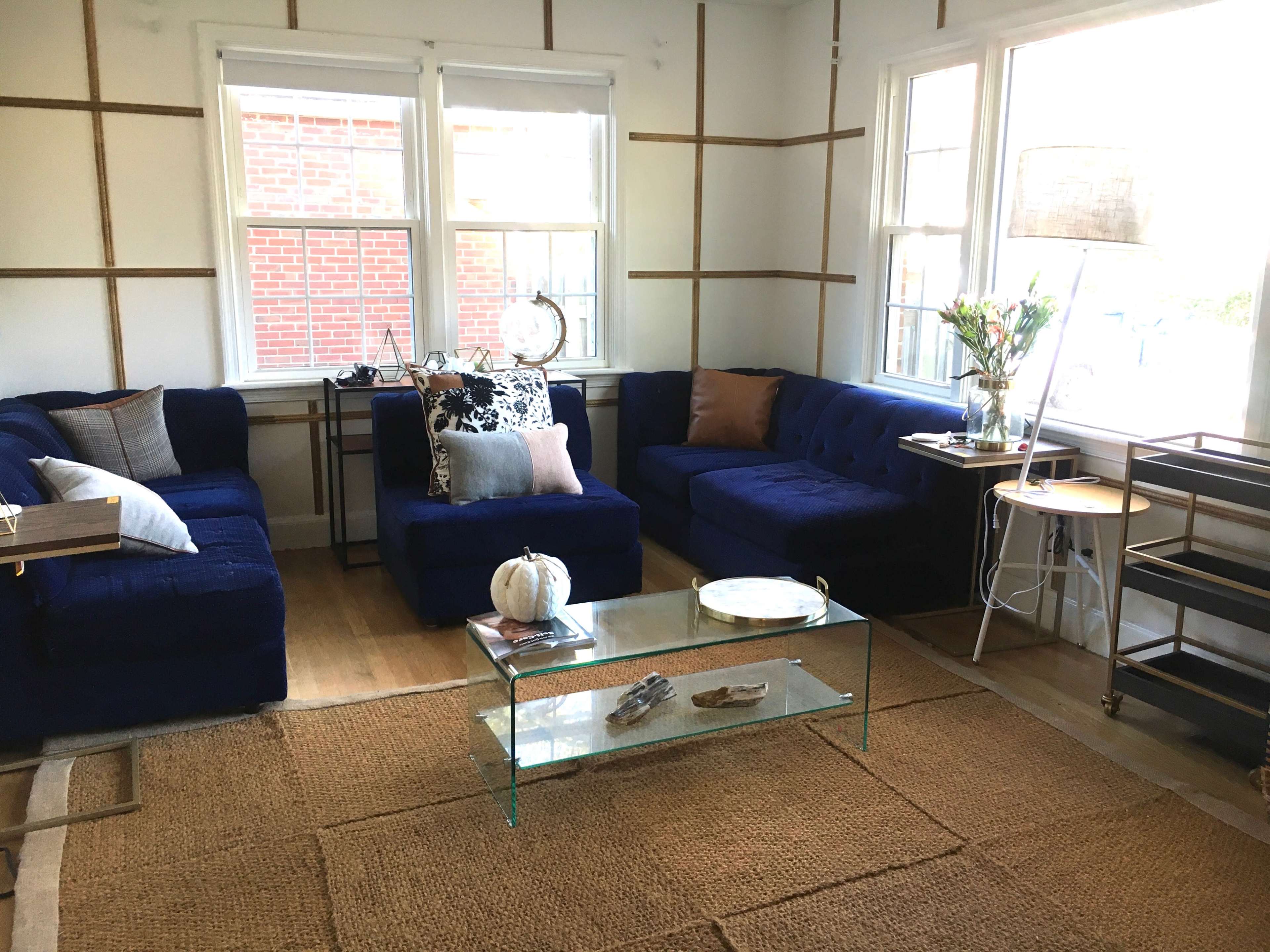 The living room features blue sofas arranged around a glass coffee table, with large windows allowing natural light and a jute rug on the wooden floor.