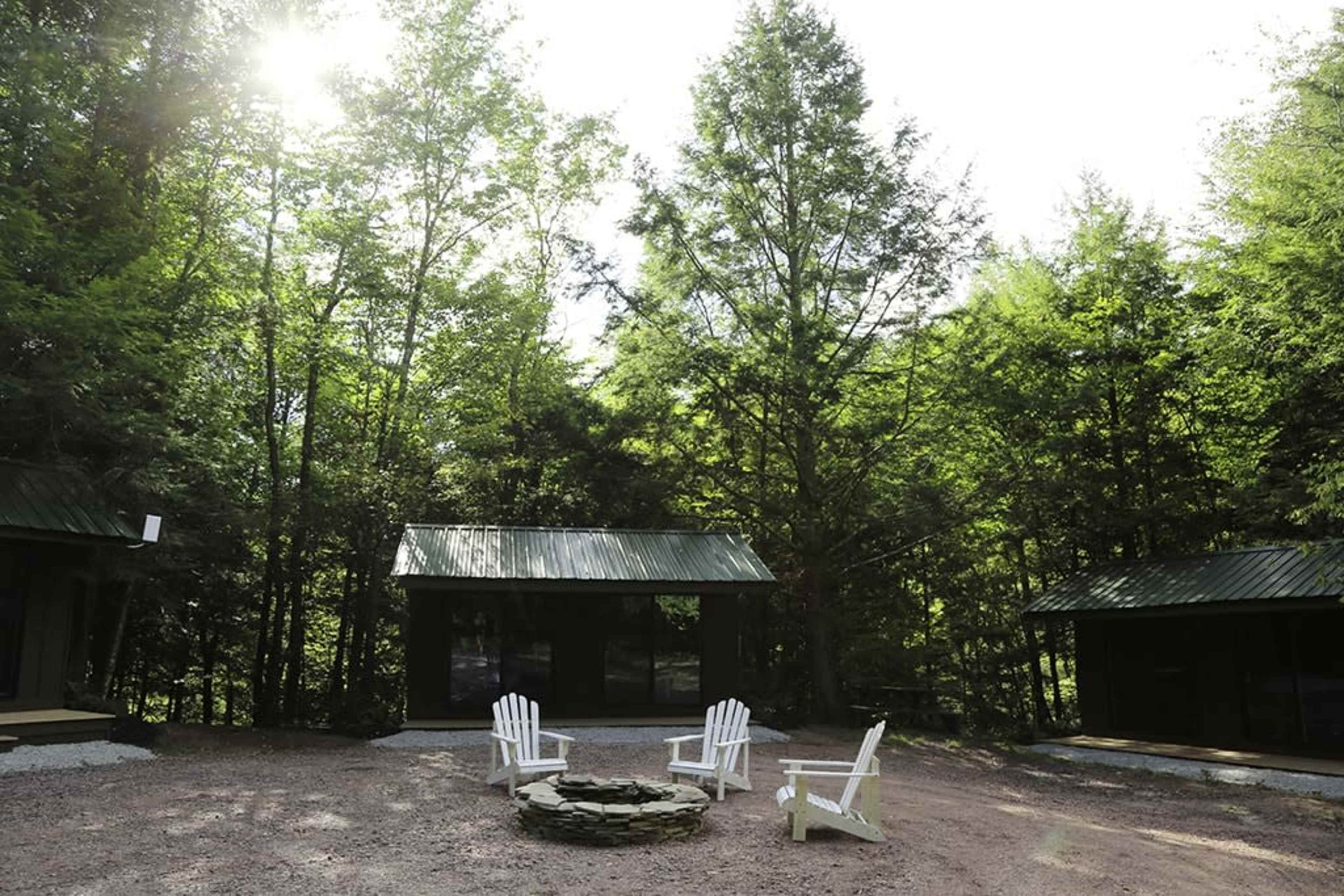 A circular stone fire pit surrounded by four white Adirondack chairs, with a backdrop of green trees and two cabins.