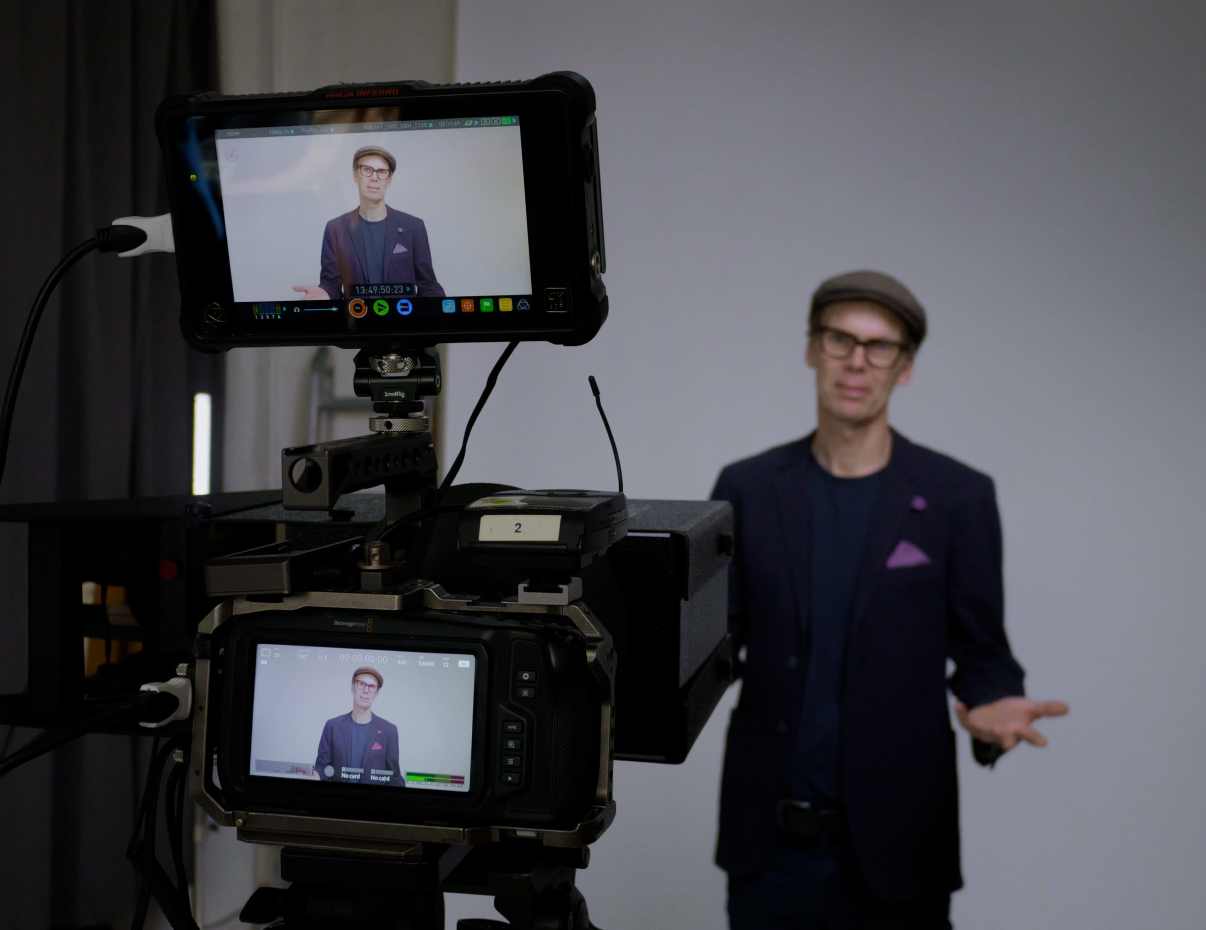 A speaker wearing a blazer and a flat cap gestures while being filmed by a camera setup in a studio.