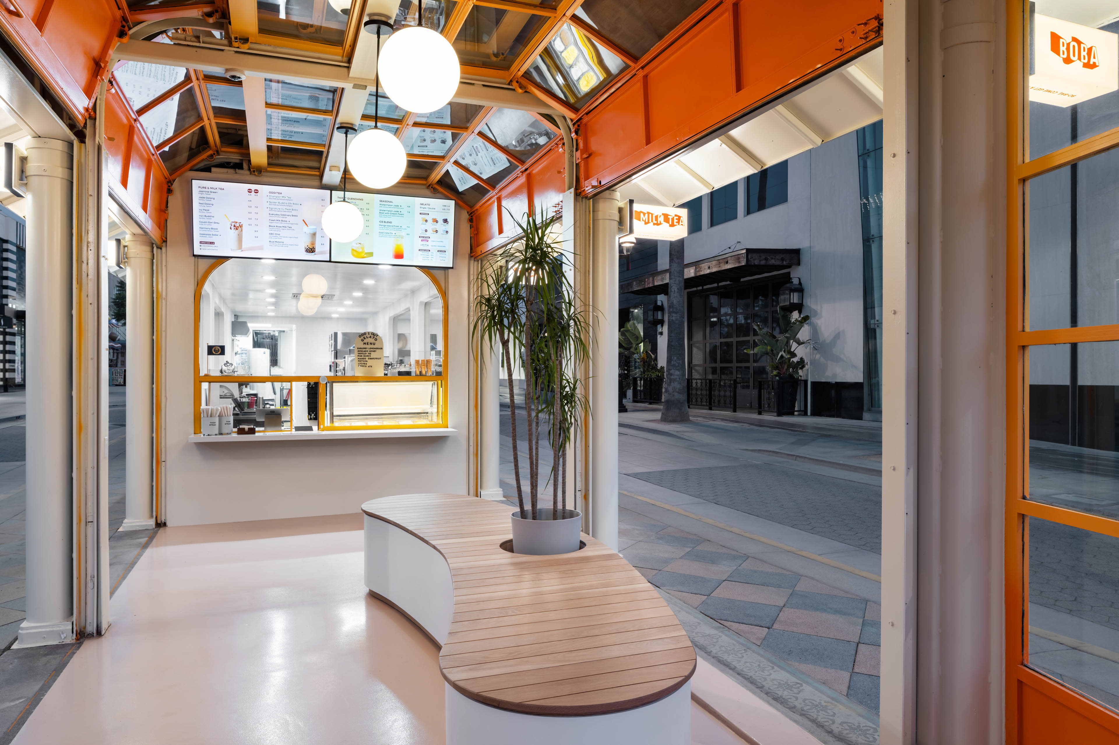 The image shows a brightly lit kiosk with a curved wooden bench, large overhead lights, and digital menus displayed on screens.