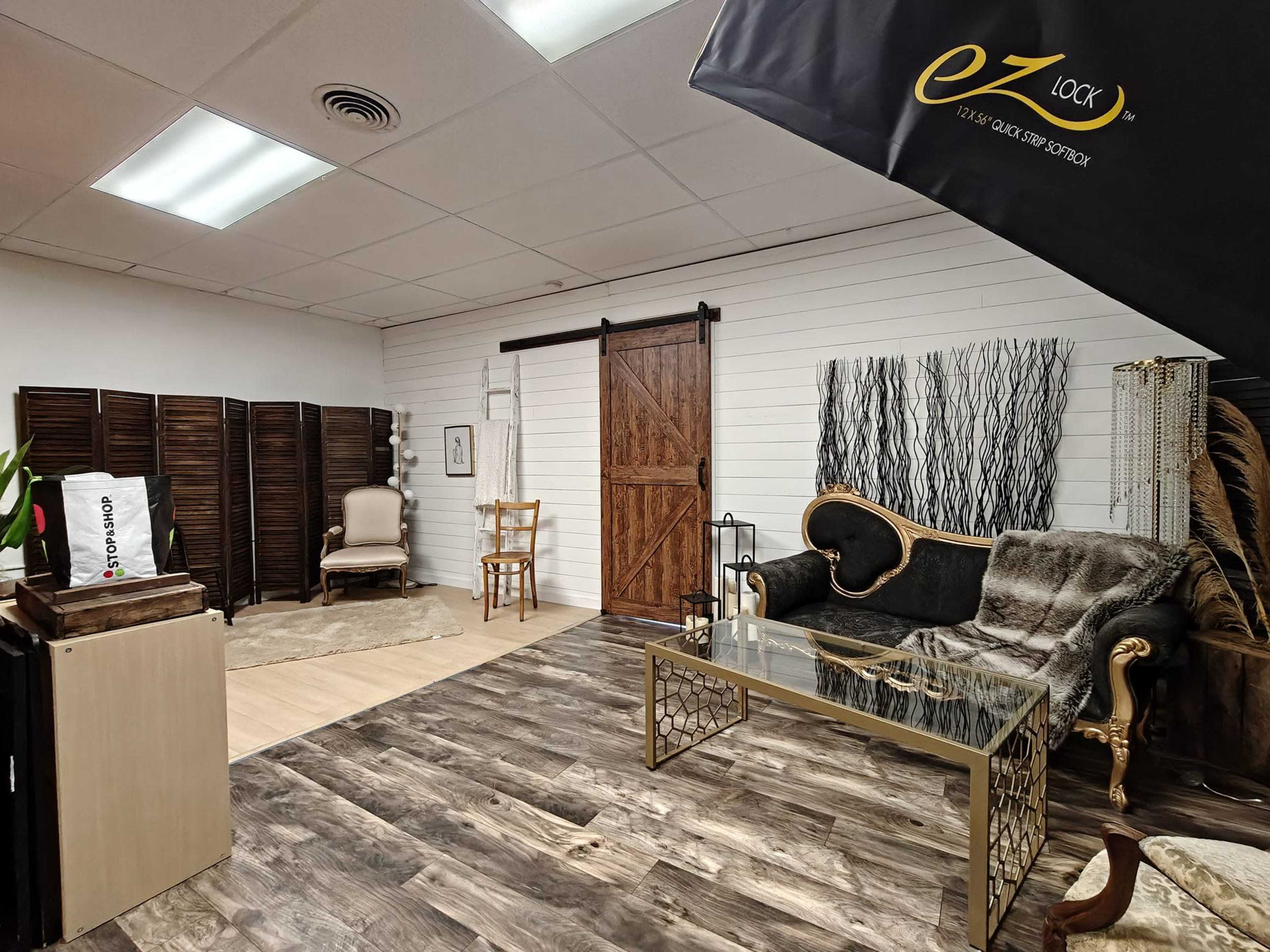 The image shows a styled interior space featuring a mix of modern and vintage furniture, wooden accents, and decorative elements against a backdrop of wooden flooring and white paneled walls.