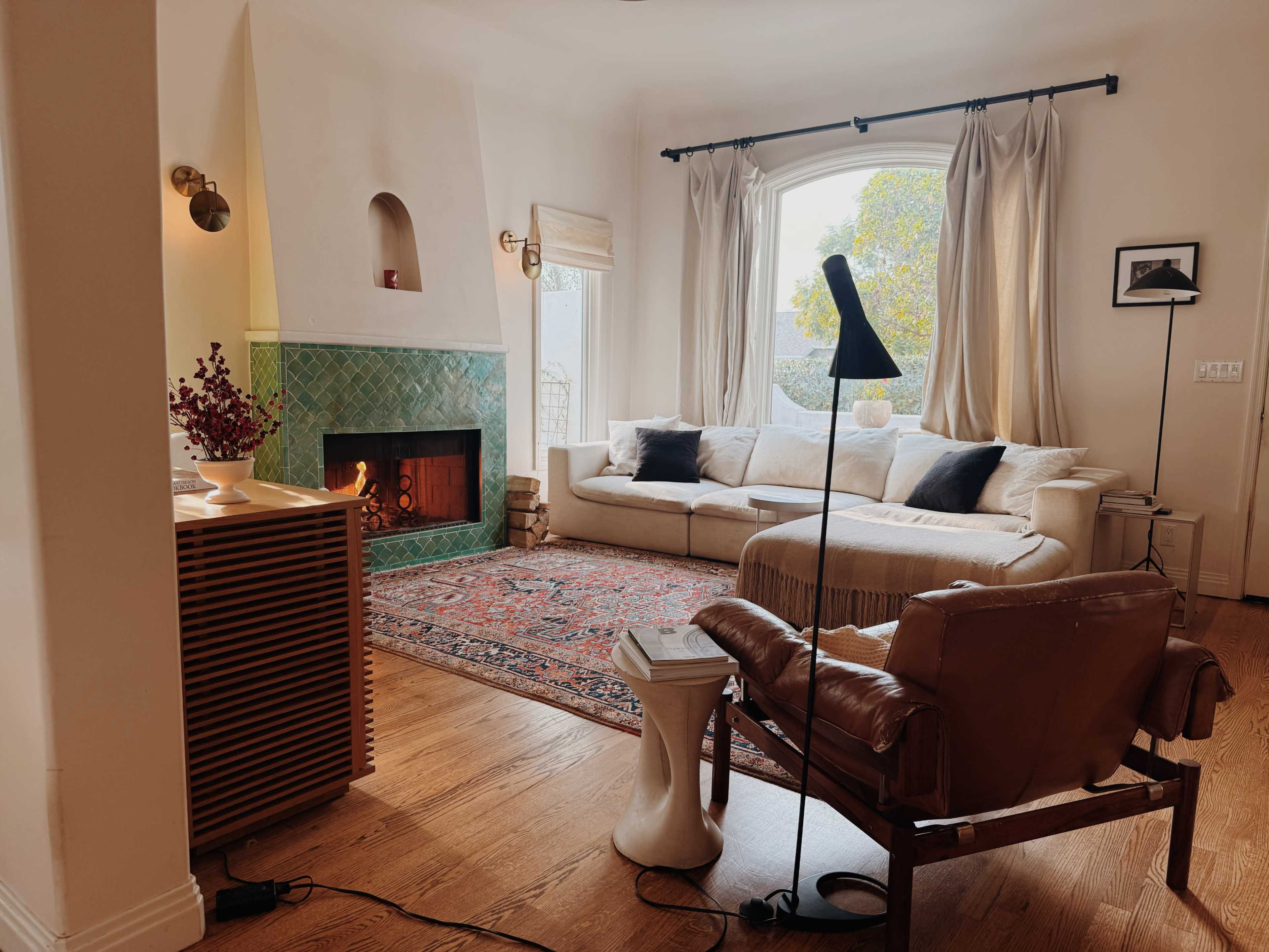 The image shows a cozy living room featuring a beige sectional sofa, a green tiled fireplace, a patterned rug, and a vintage-style lounge chair with a floor lamp beside it.